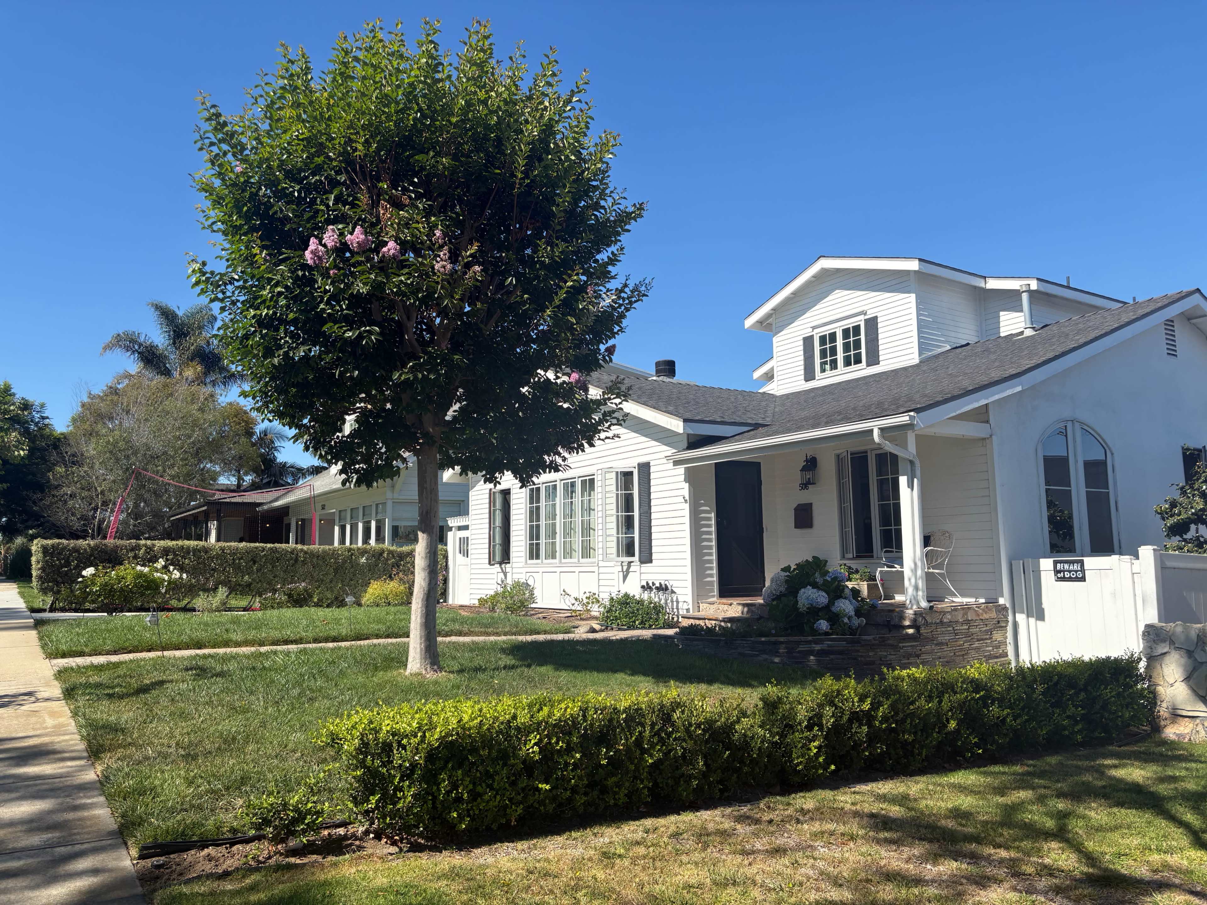 A white, single-story house with a well-manicured lawn and a large tree stands on a sunny street.