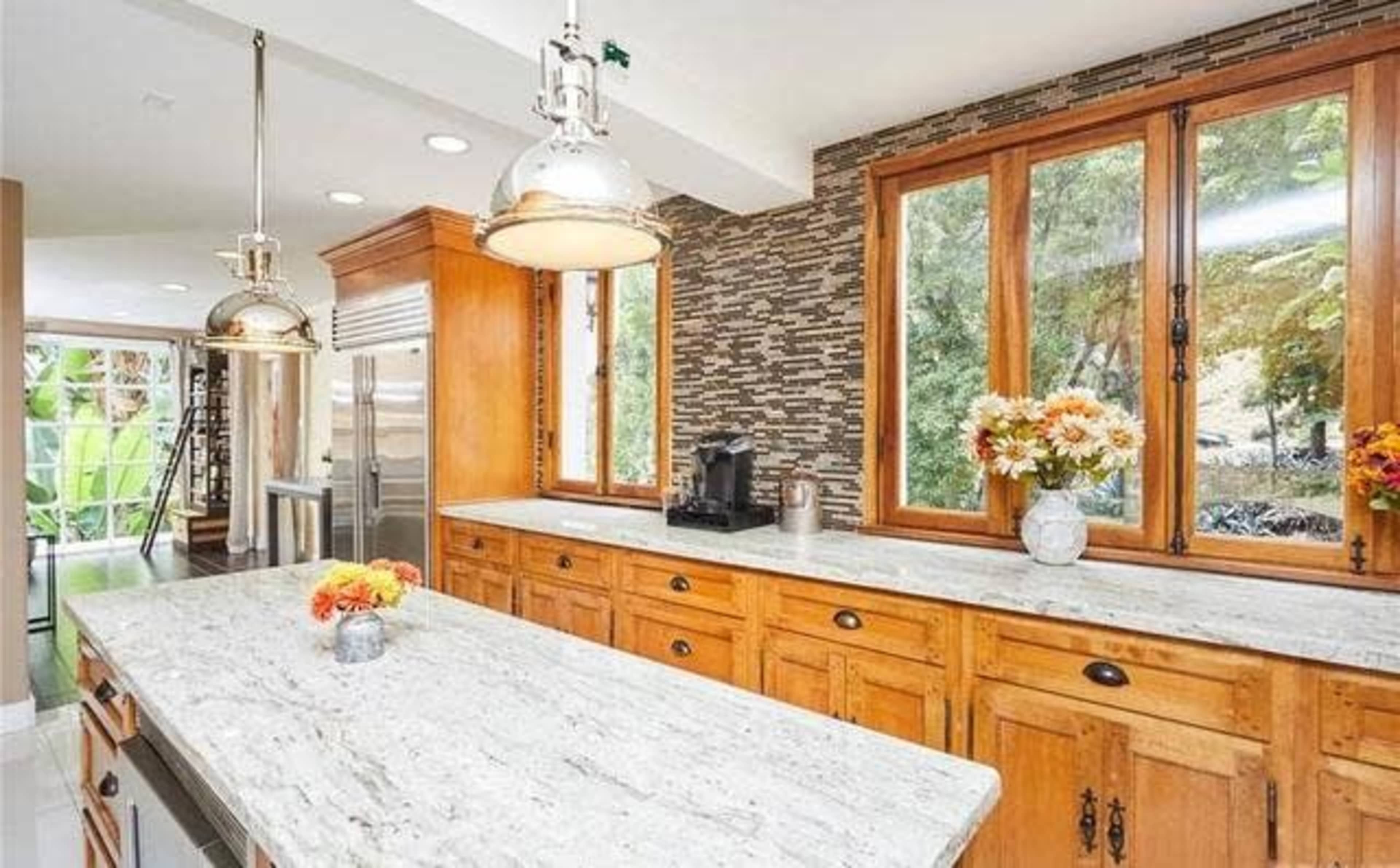 The kitchen features a large granite island with wooden cabinetry and large windows overlooking greenery.