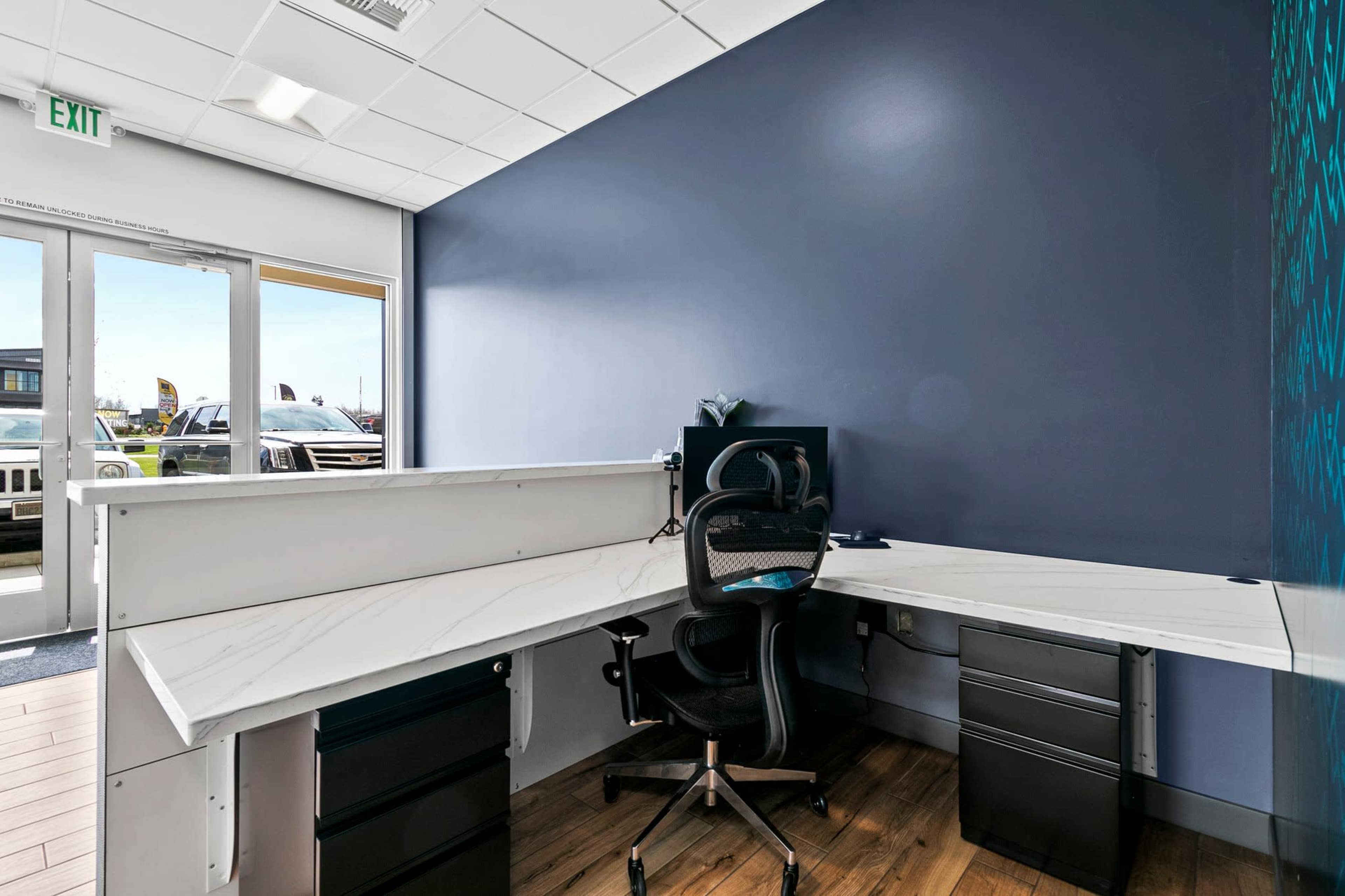 The image shows a modern office workspace featuring a corner desk with a black ergonomic chair, against a dark blue wall, and a view of an exit door with natural light coming in.