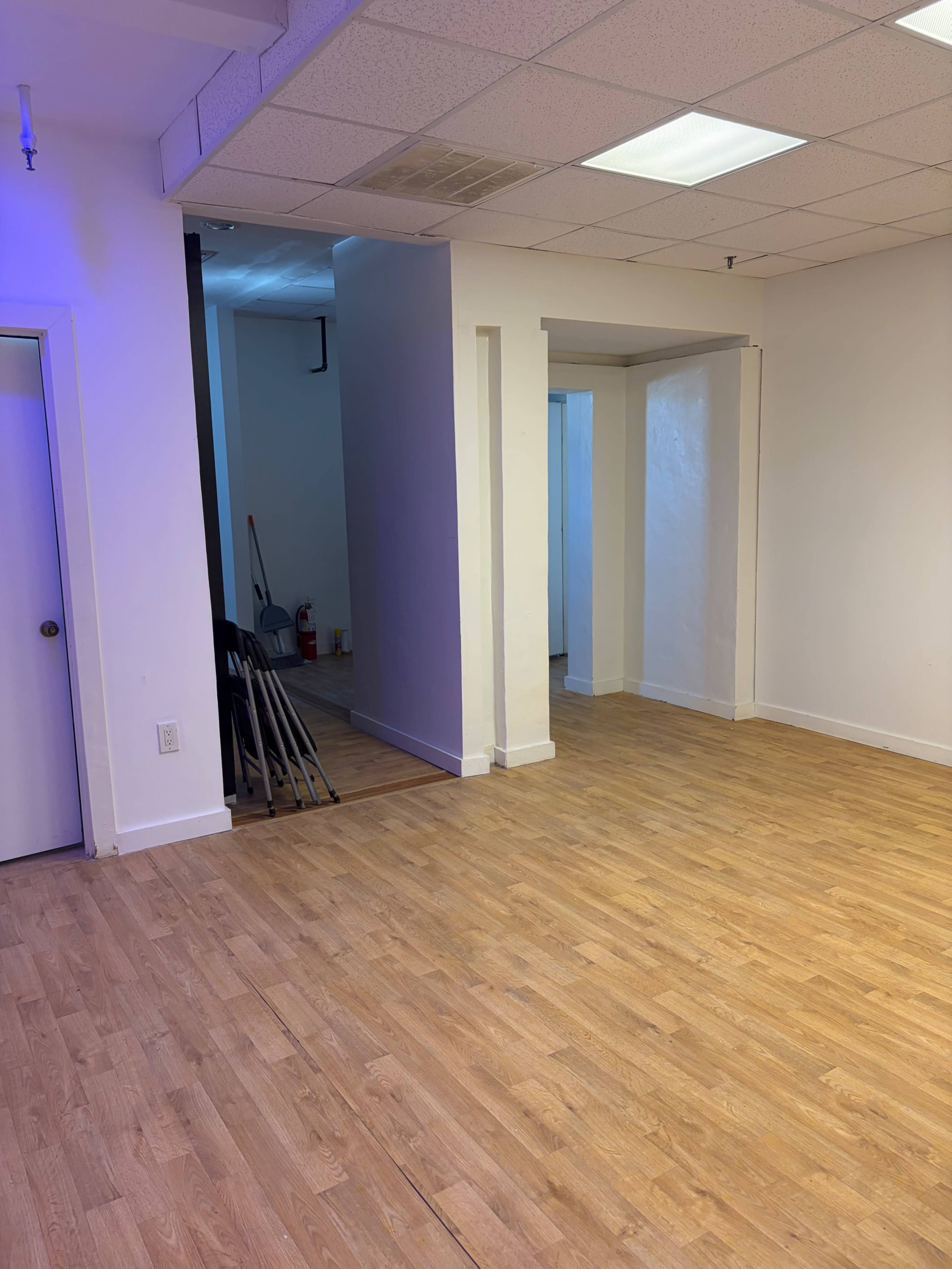 The image shows a spacious, empty room with wooden flooring, white walls, and a few doors leading to adjacent areas.