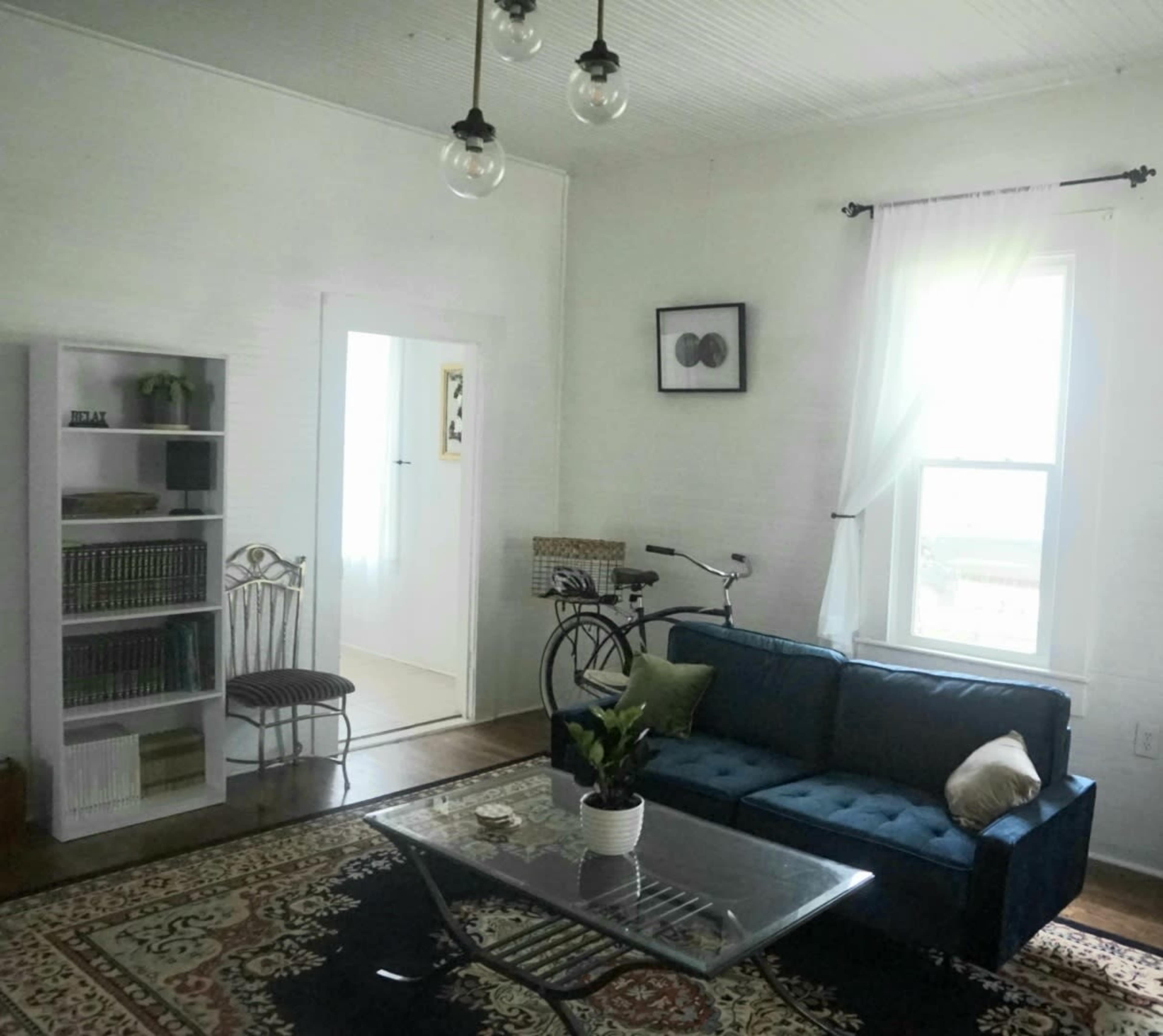 A modern living room features a blue sofa, a glass coffee table, a bookshelf, and a bicycle next to a window.