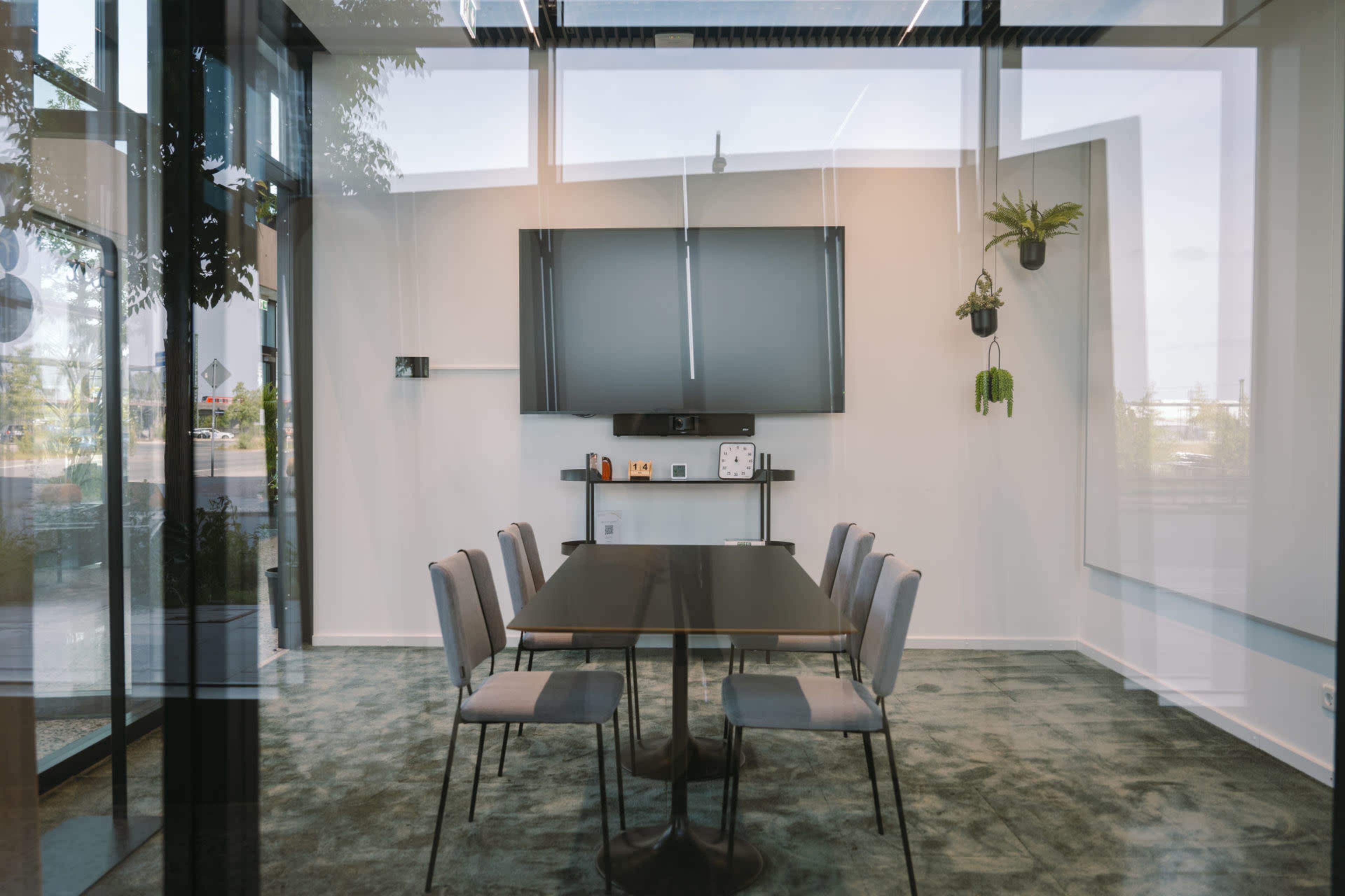 The image shows a modern meeting room with a rectangular table and six chairs, featuring a large screen mounted on the wall and plants for decoration.