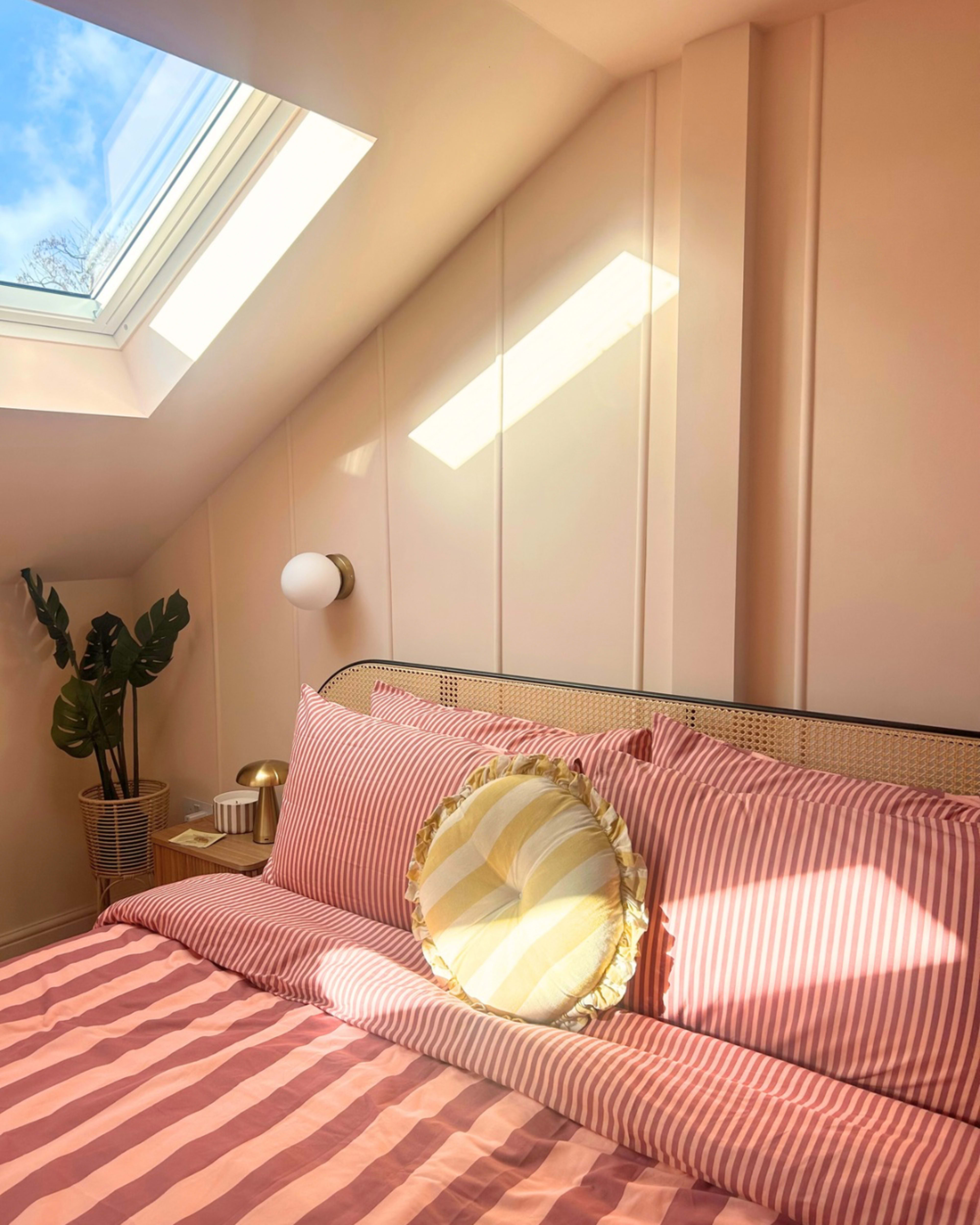 The image shows a cozy bedroom with a slanted ceiling, a striped bedspread, a round pillow, a plant in a corner, and natural light streaming through a skylight.