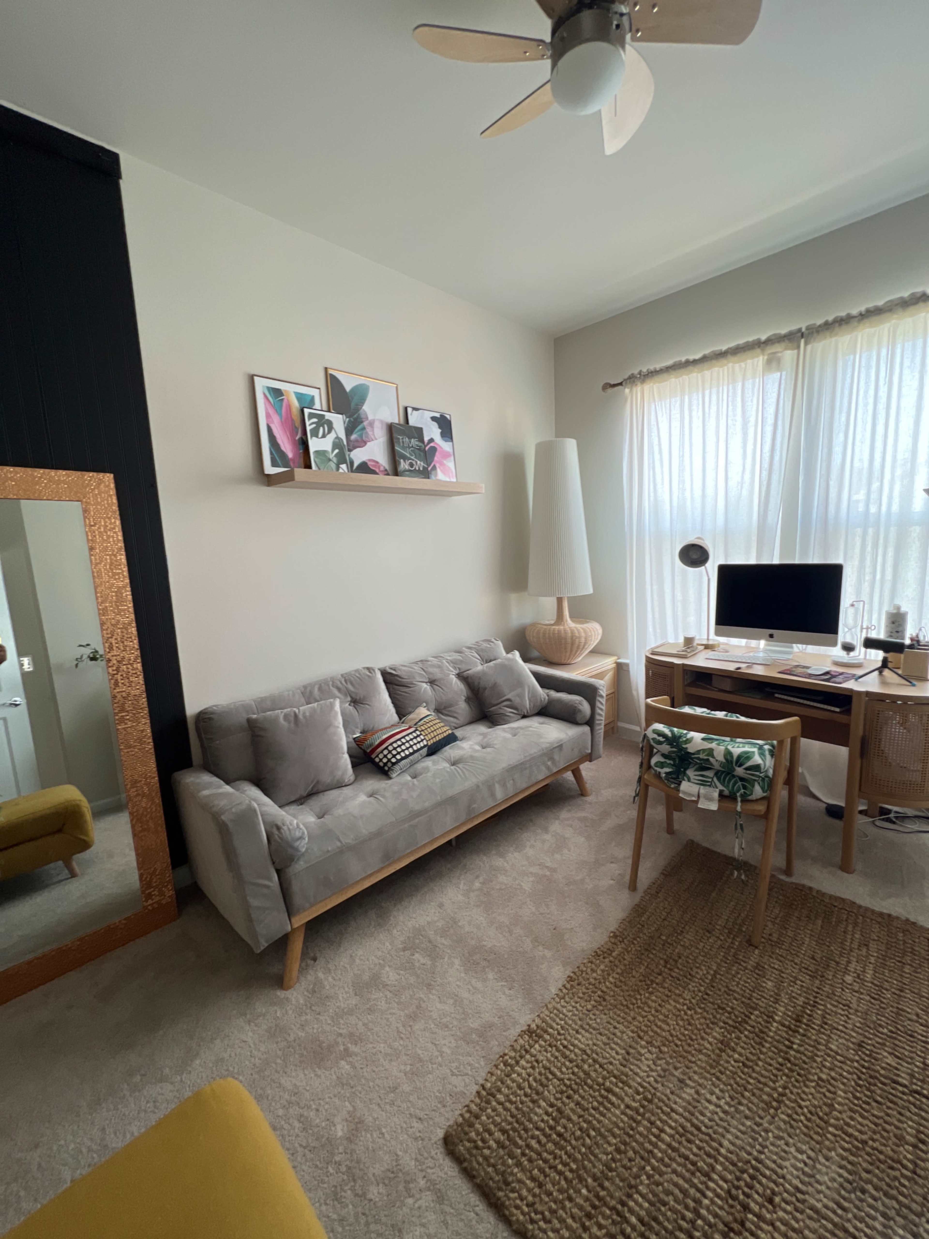 A cozy home office features a gray sofa, a wooden desk with a computer, and large windows with sheer curtains allowing natural light to fill the space.