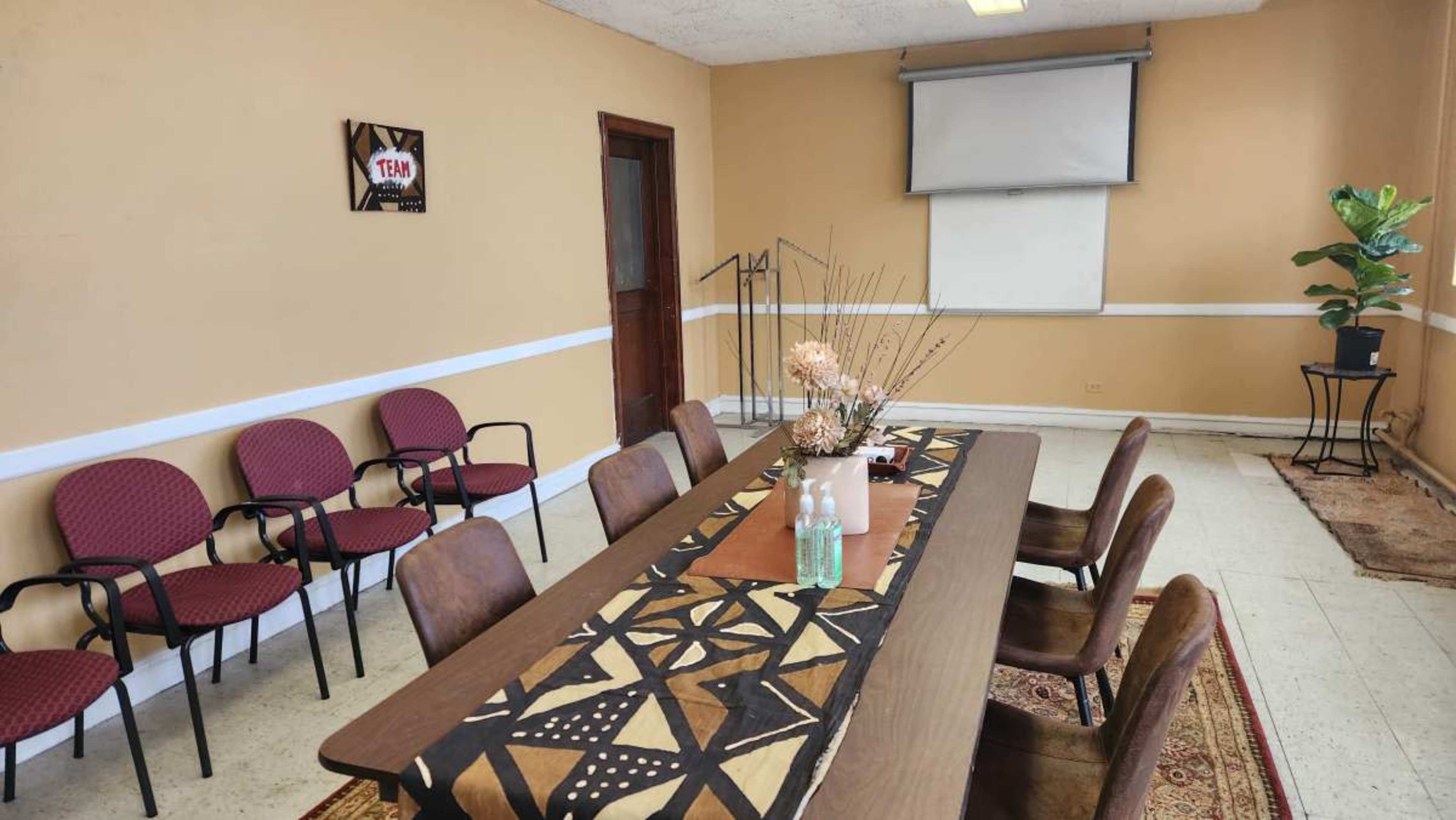 A conference room features a long table with decorative elements, surrounded by chairs, and a projector screen mounted on the wall.