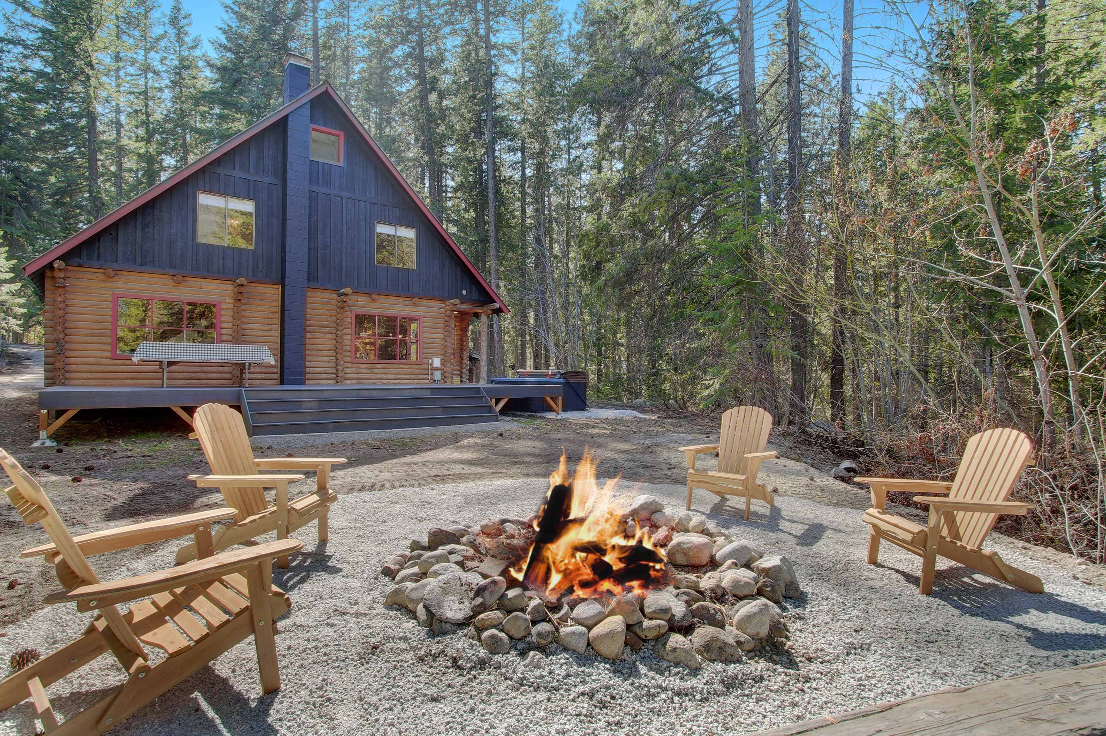 A log cabin with a red roof is set among tall trees, featuring a fire pit surrounded by wooden chairs in front of it.