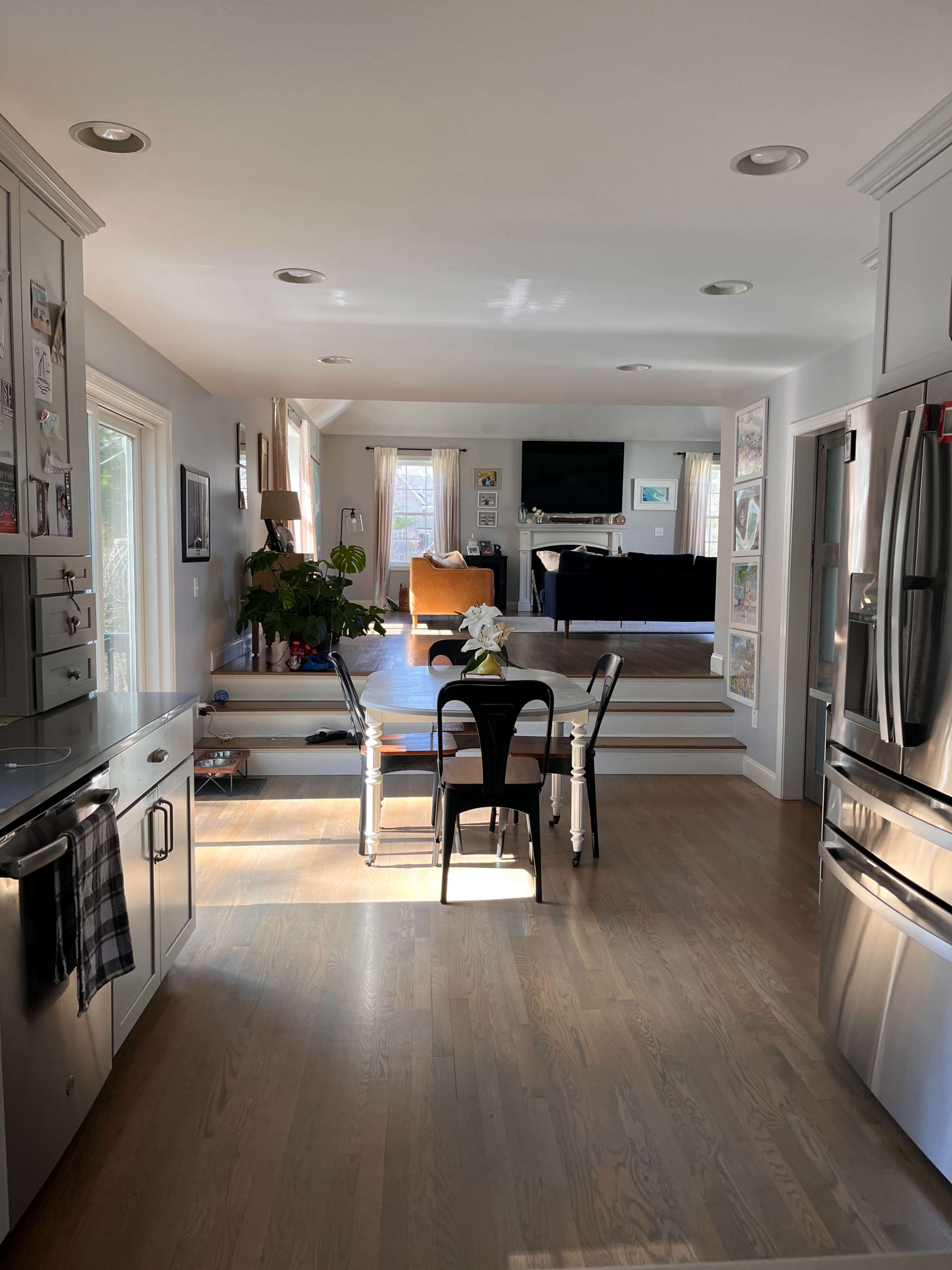 A kitchen with a dining table and chairs in the foreground, leading to an open living area with large windows and a television.