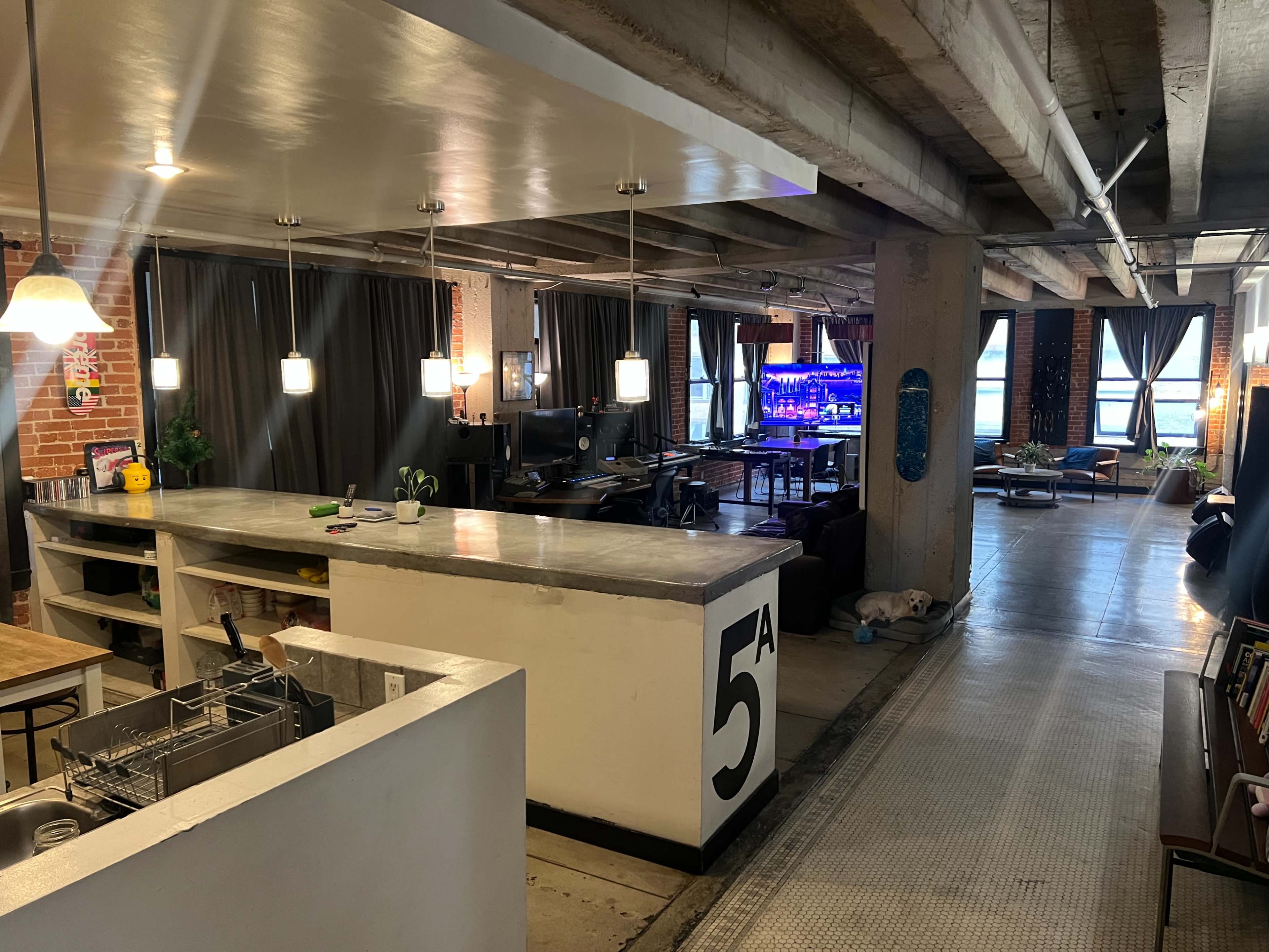 The image shows an open, modern space with a kitchen area in the foreground and a coworking area featuring desks and a large screen in the background.
