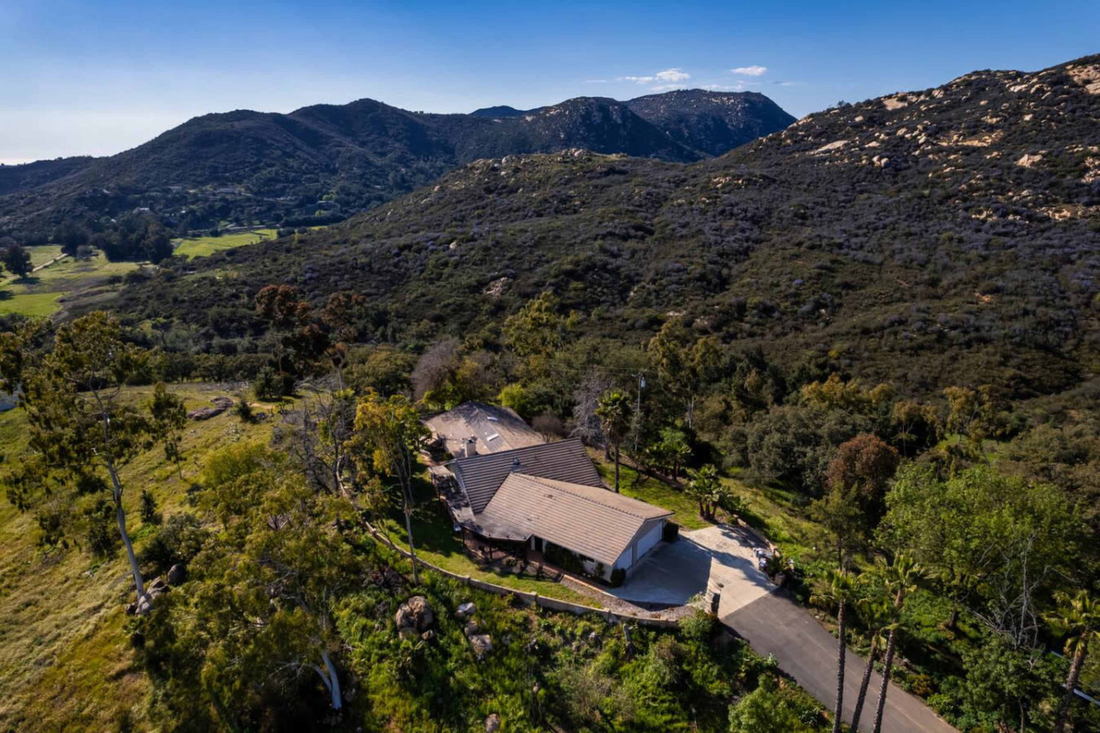 A modern house is situated on a hillside surrounded by green trees and rolling mountains.