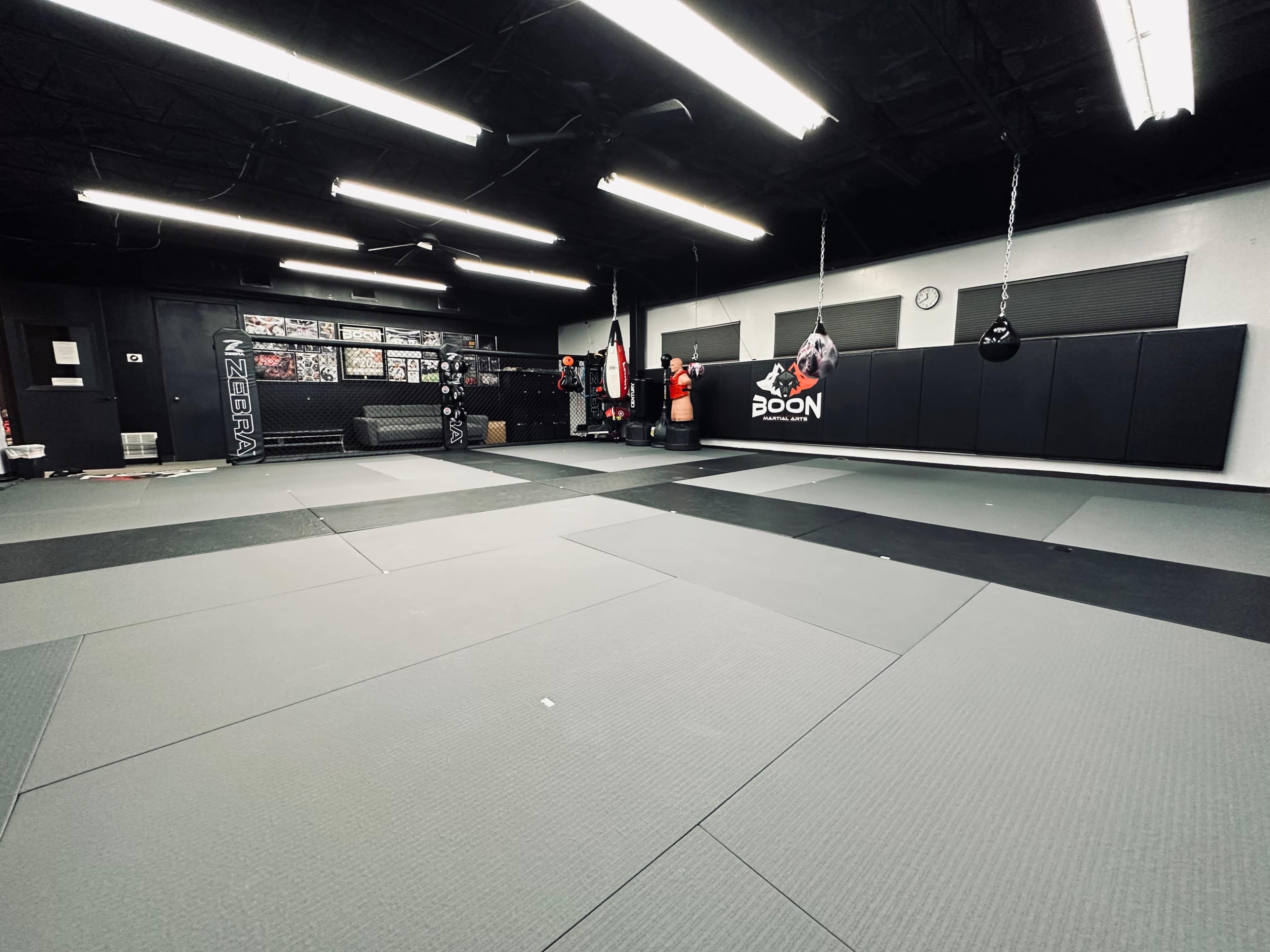 A training facility with gray mats, black wall padding, and several training equipment, including heavy bags and boxing gloves.
