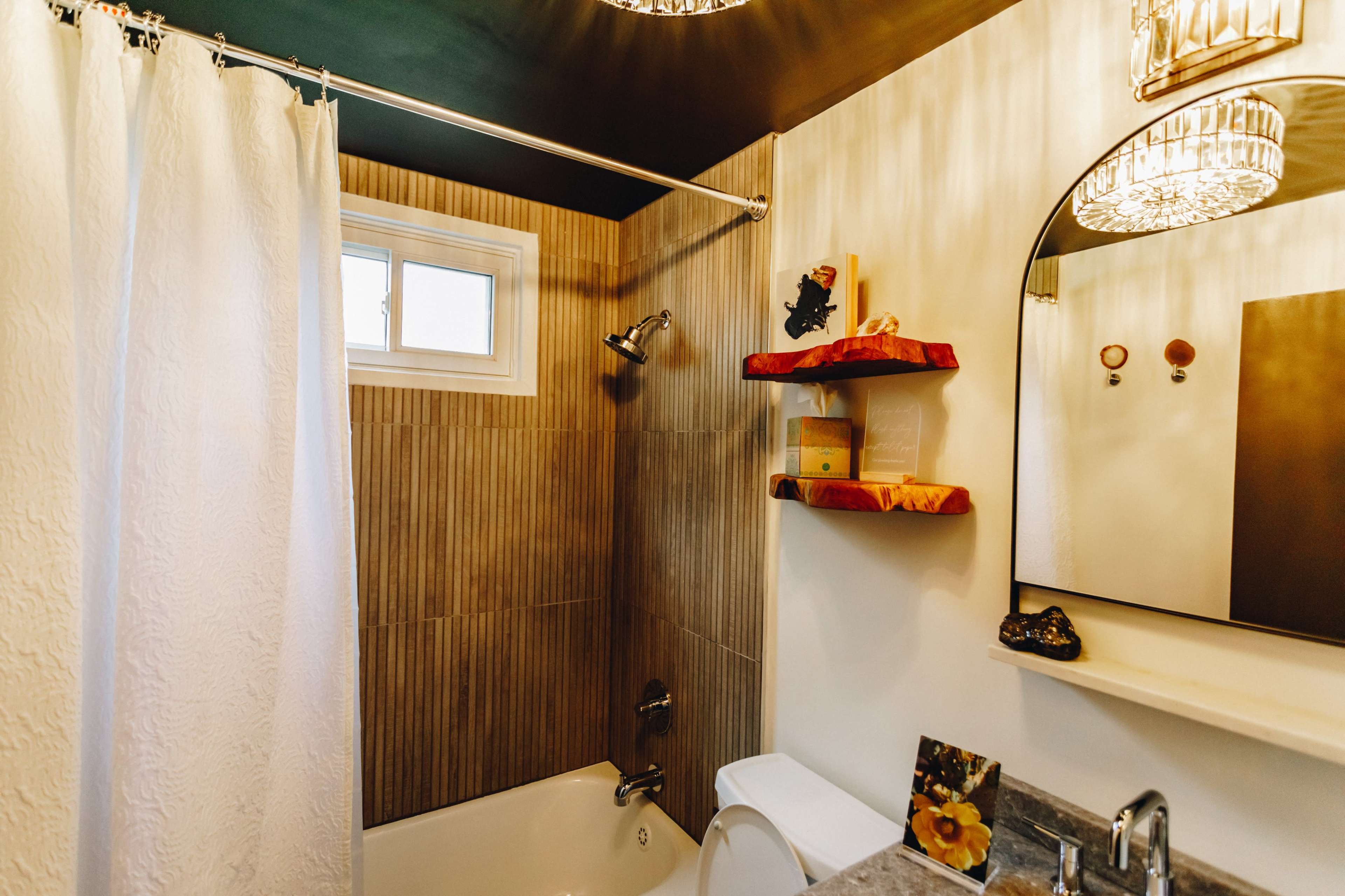 A compact bathroom features a bathtub with a shower, a window, decorative shelves, and a round mirror with two light fixtures above.