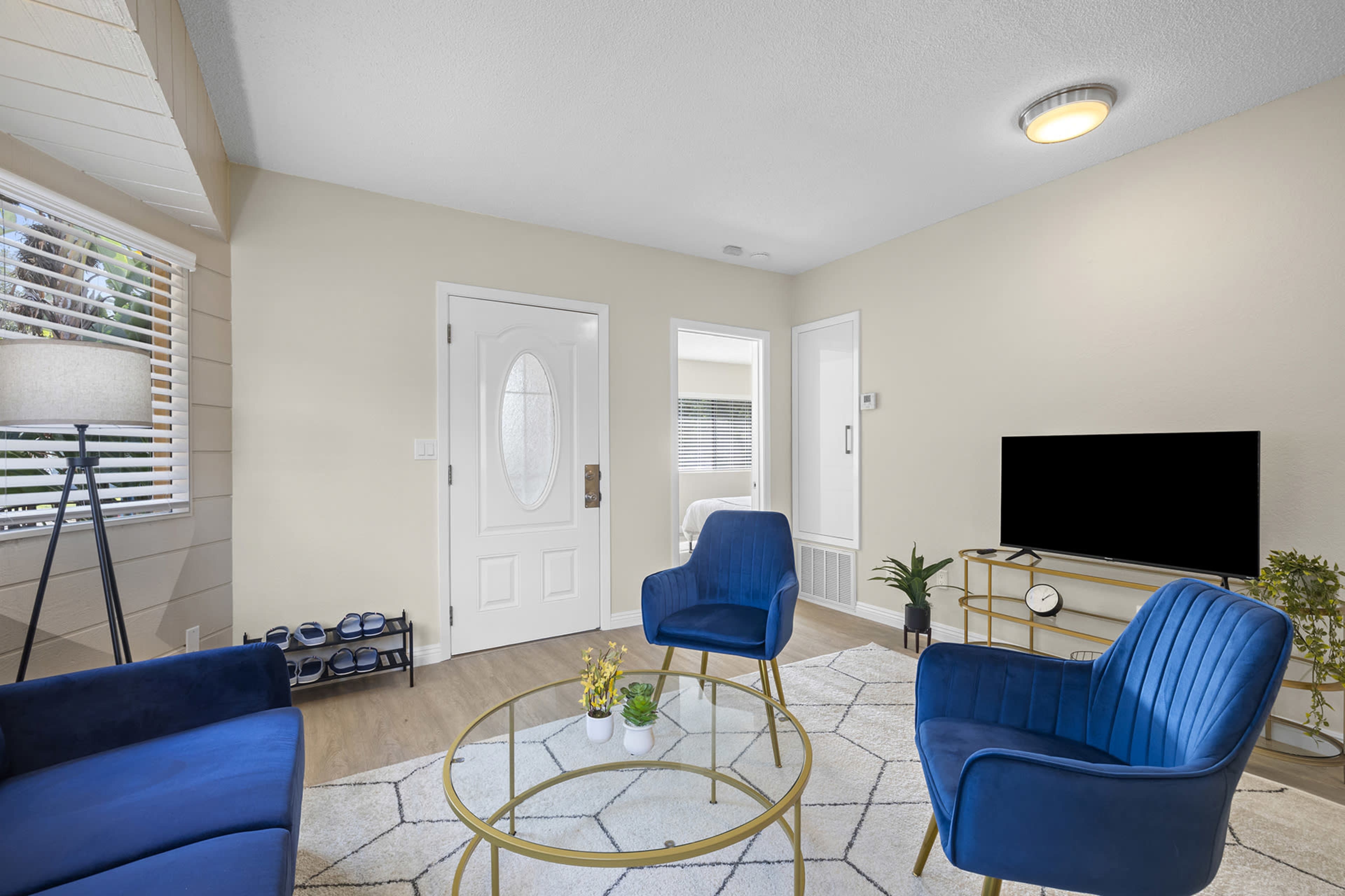 The room features two blue chairs, a glass coffee table, a television on a stand, and a front door, with windows allowing natural light.