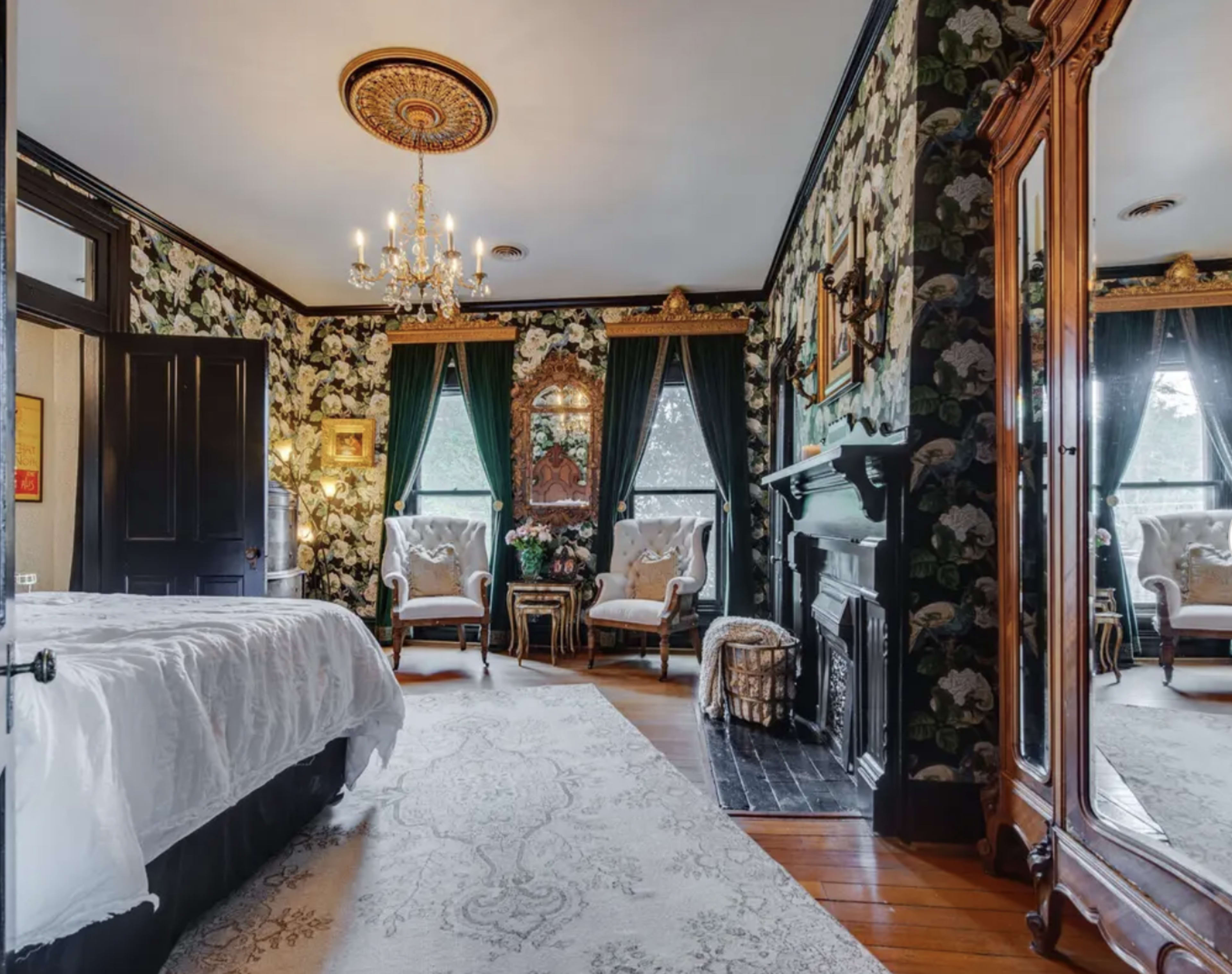 A furnished bedroom with floral wallpaper, a fireplace, and two upholstered chairs near a large window.