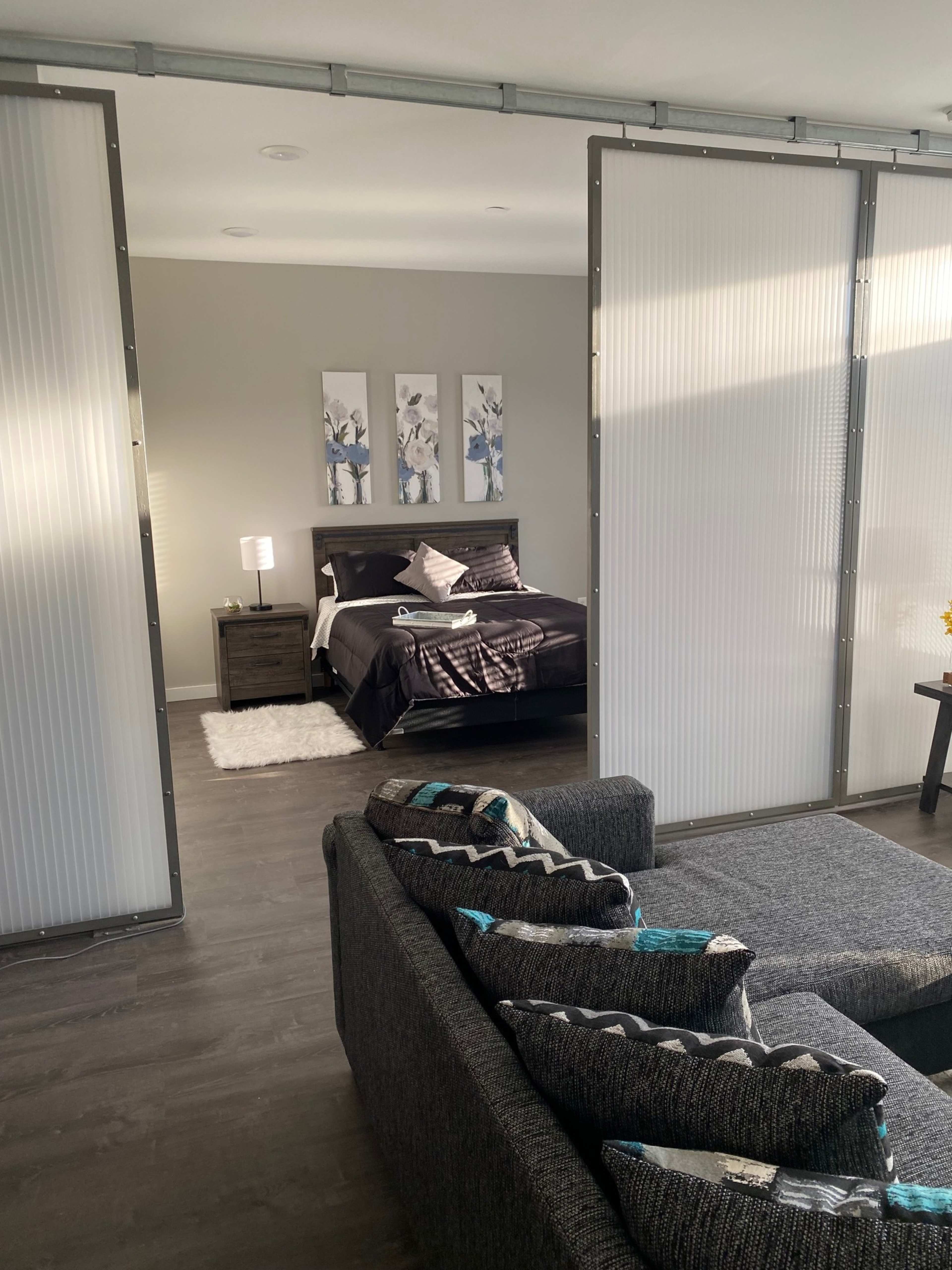 A modern living space with a seating area featuring a sectional couch and a bedroom partially separated by sliding panels that include a bed and nightstand.