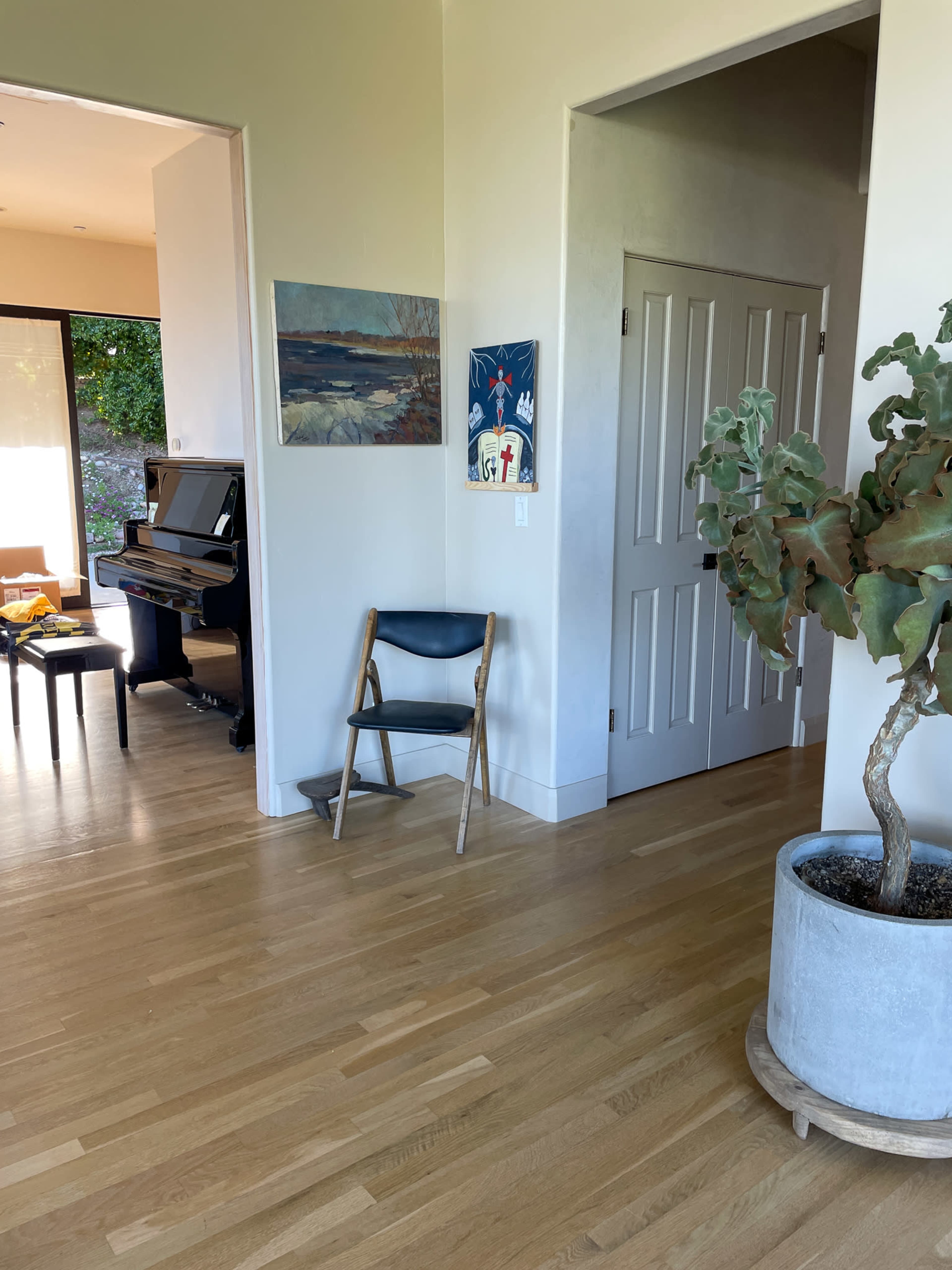 A bright, open hallway features a wooden floor, a small plant in a large pot, a folding chair, and a doorway leading to a room with a piano.