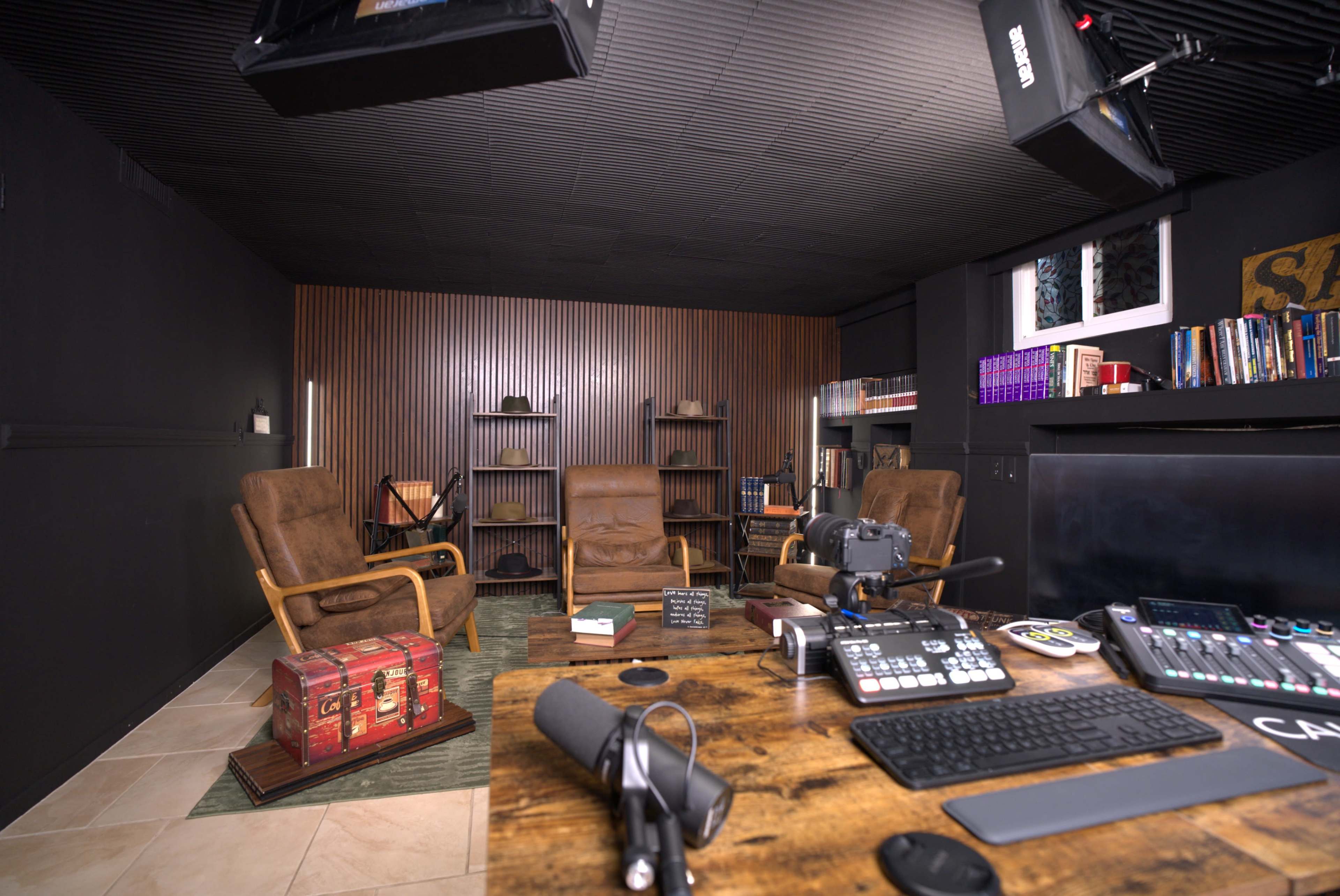 The image depicts a modern audio-visual recording studio featuring two brown armchairs, a wooden desk with recording equipment, and shelves filled with books against a dark, paneled wall.