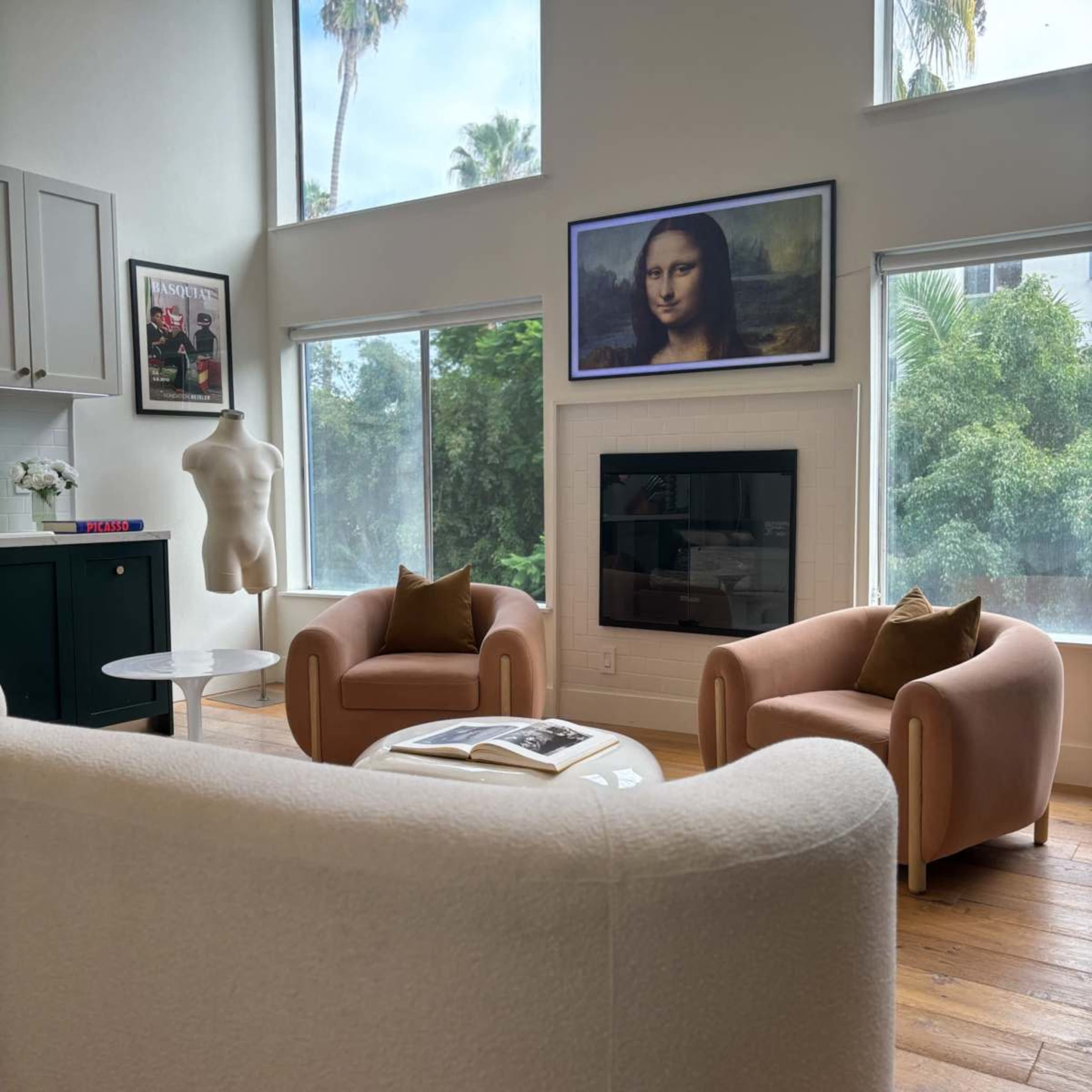 A modern living room with two pink armchairs, a mannequin, a coffee table, and a framed portrait of the Mona Lisa above a fireplace, surrounded by large windows.