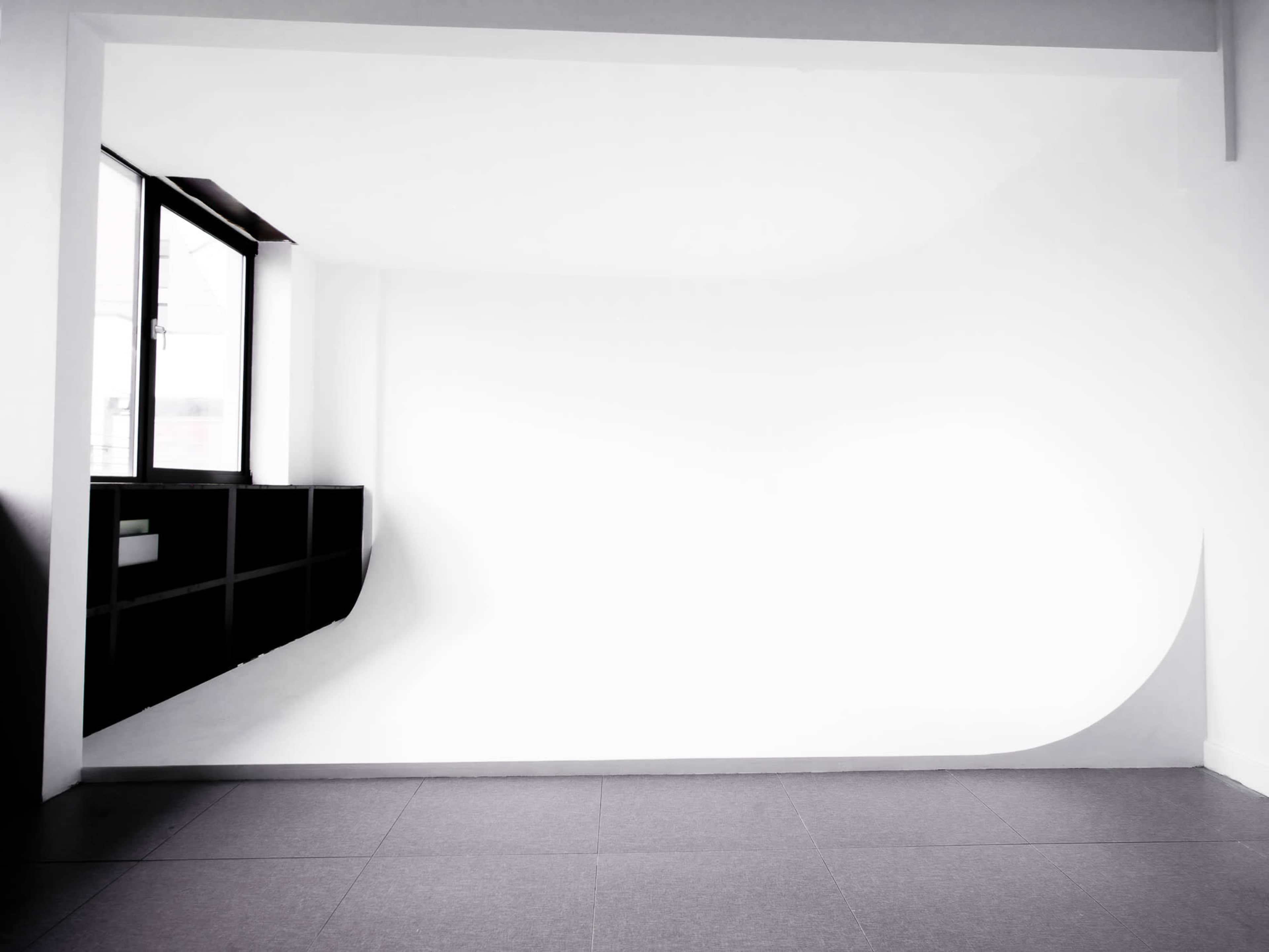 The image shows a minimalistic studio with a curved white backdrop and a large window on one side.