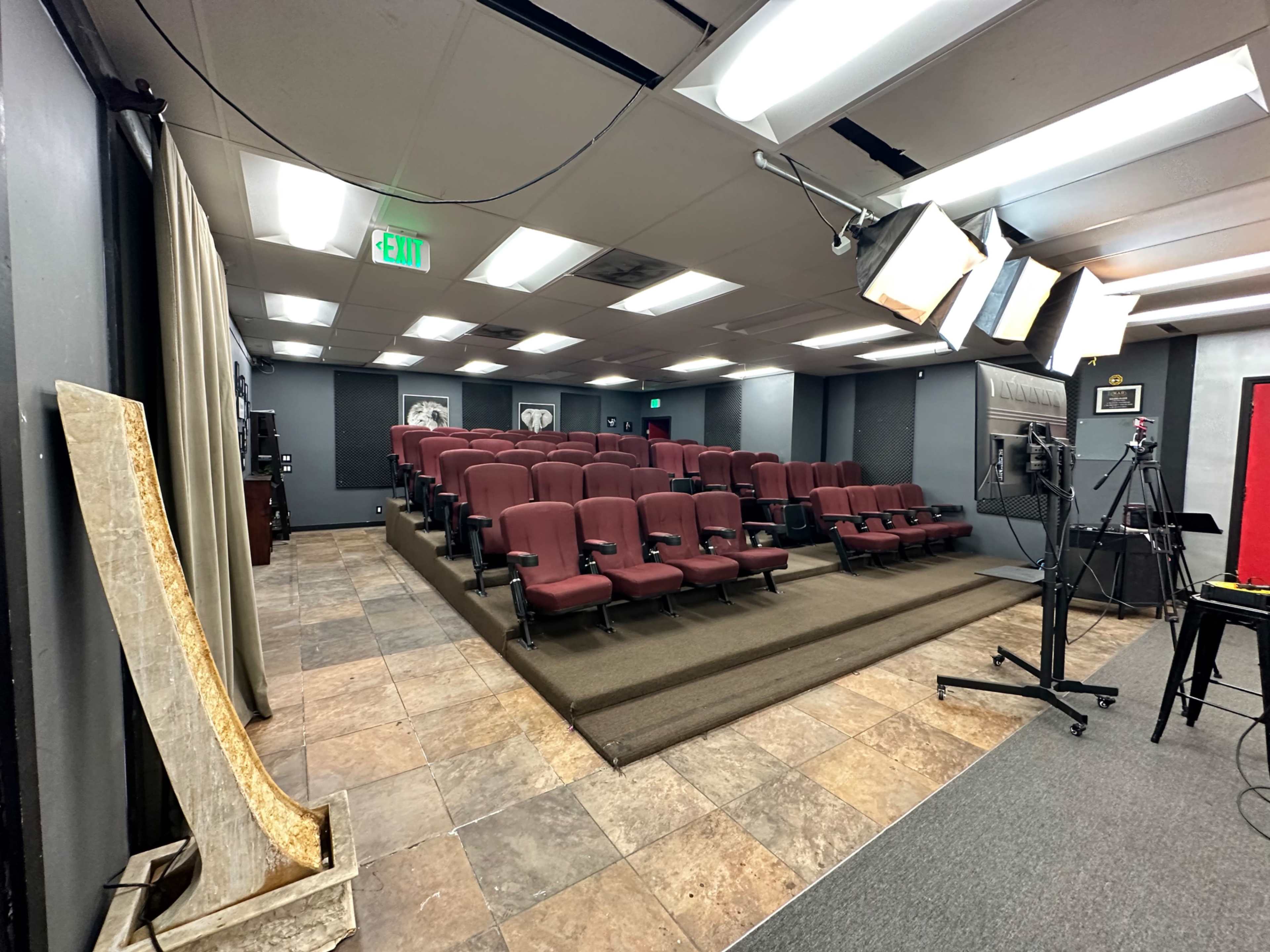 A small auditorium with rows of burgundy seating, a stage setup with lighting equipment, and a tiled floor.