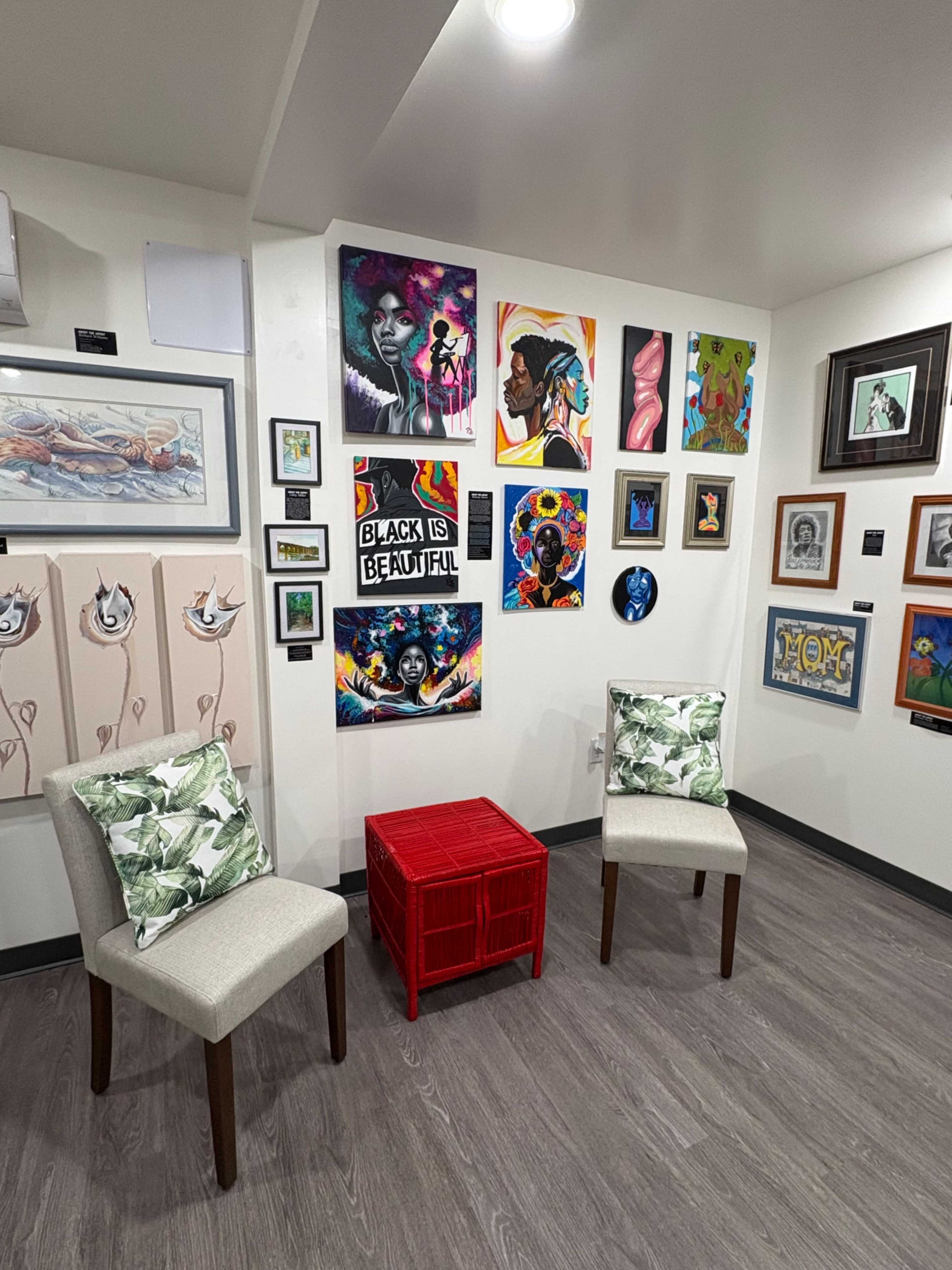 The image shows an art gallery wall adorned with various framed artworks, alongside two chairs and a red crate in a corner.