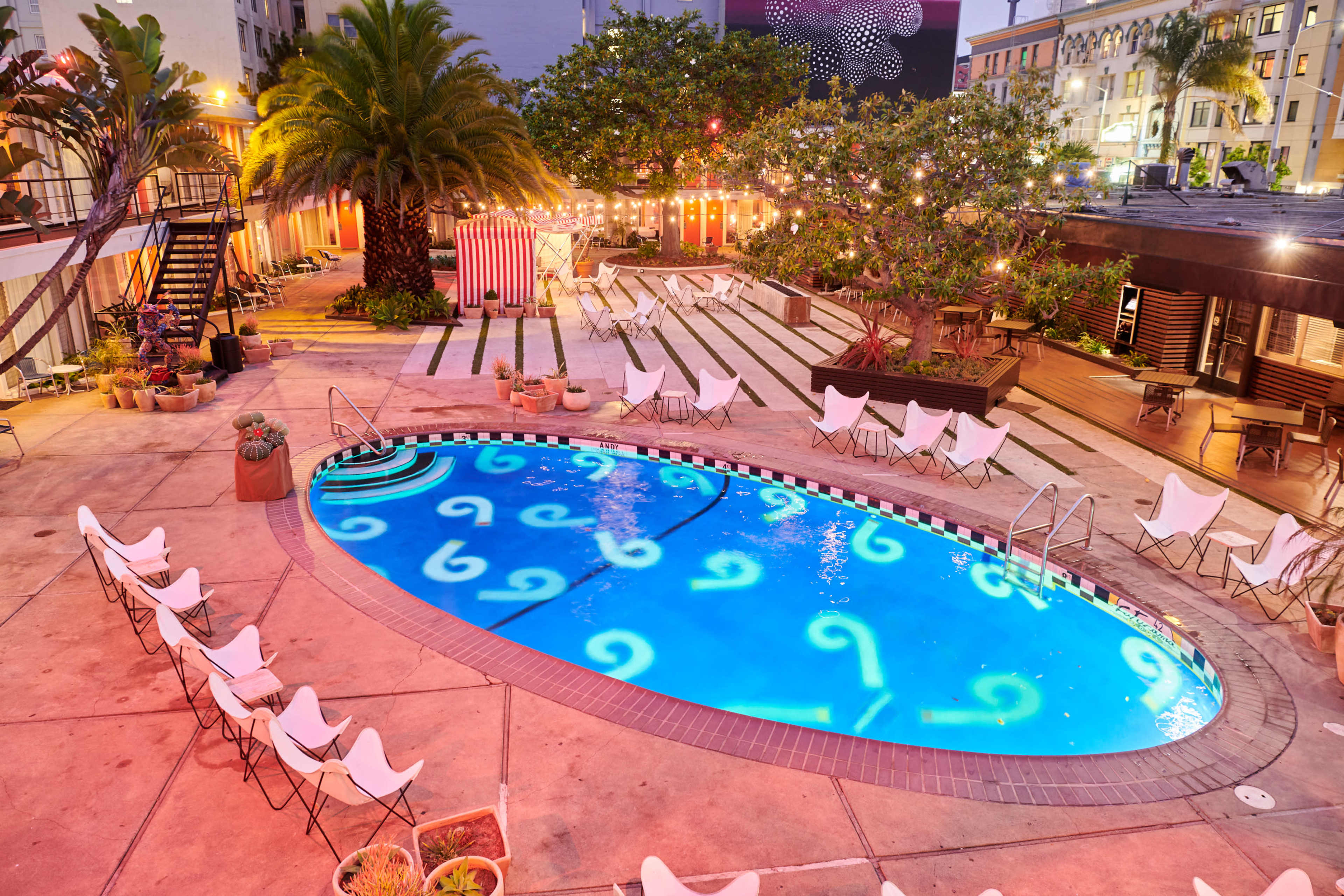 A diagonal view of a pool surrounded by lounge chairs, palm trees, and decorative string lights in an urban outdoor setting.