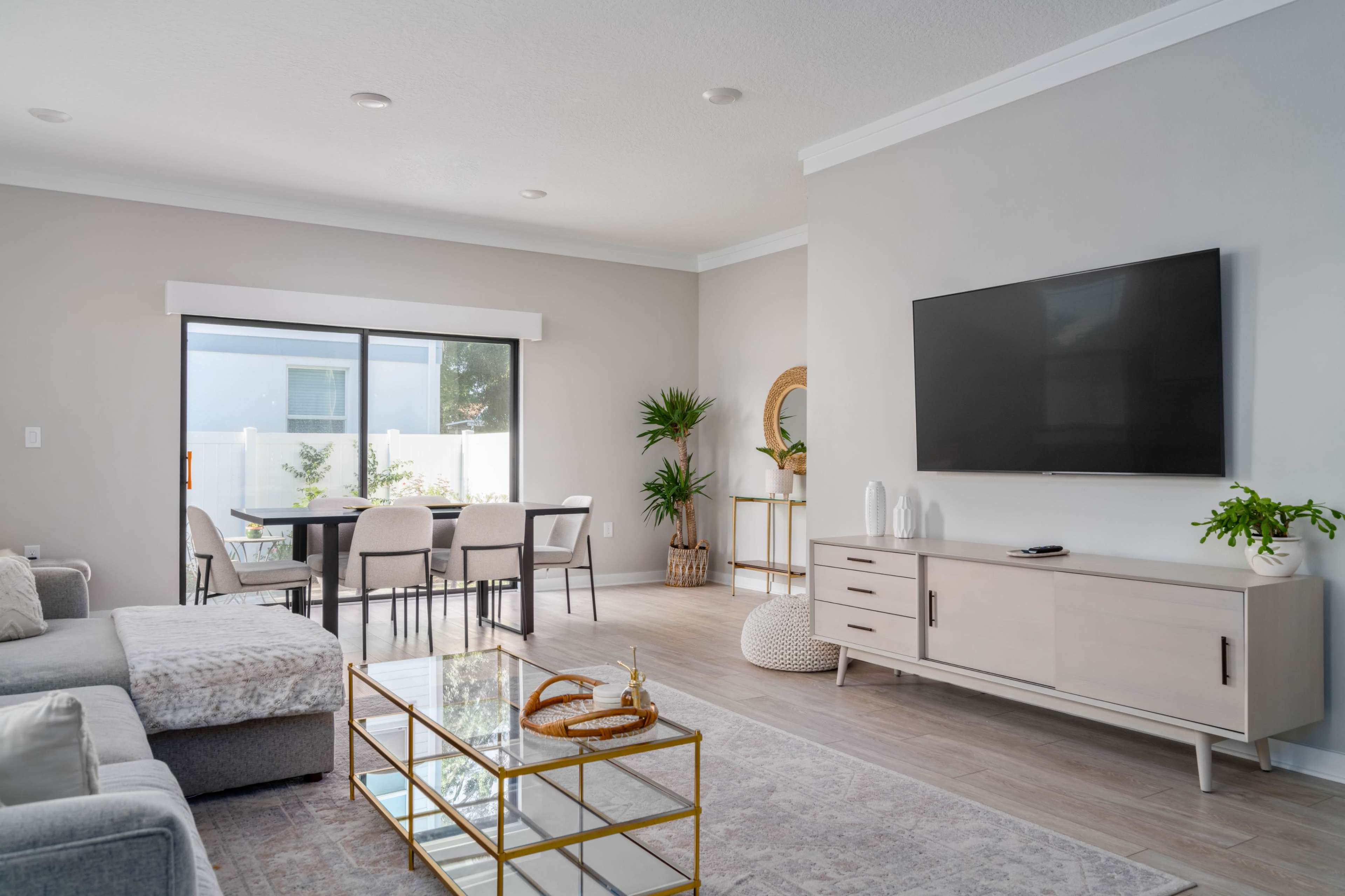 A modern living room with a gray sectional sofa, a coffee table with a gold frame, a wall-mounted television, and a dining area visible through a sliding glass door.