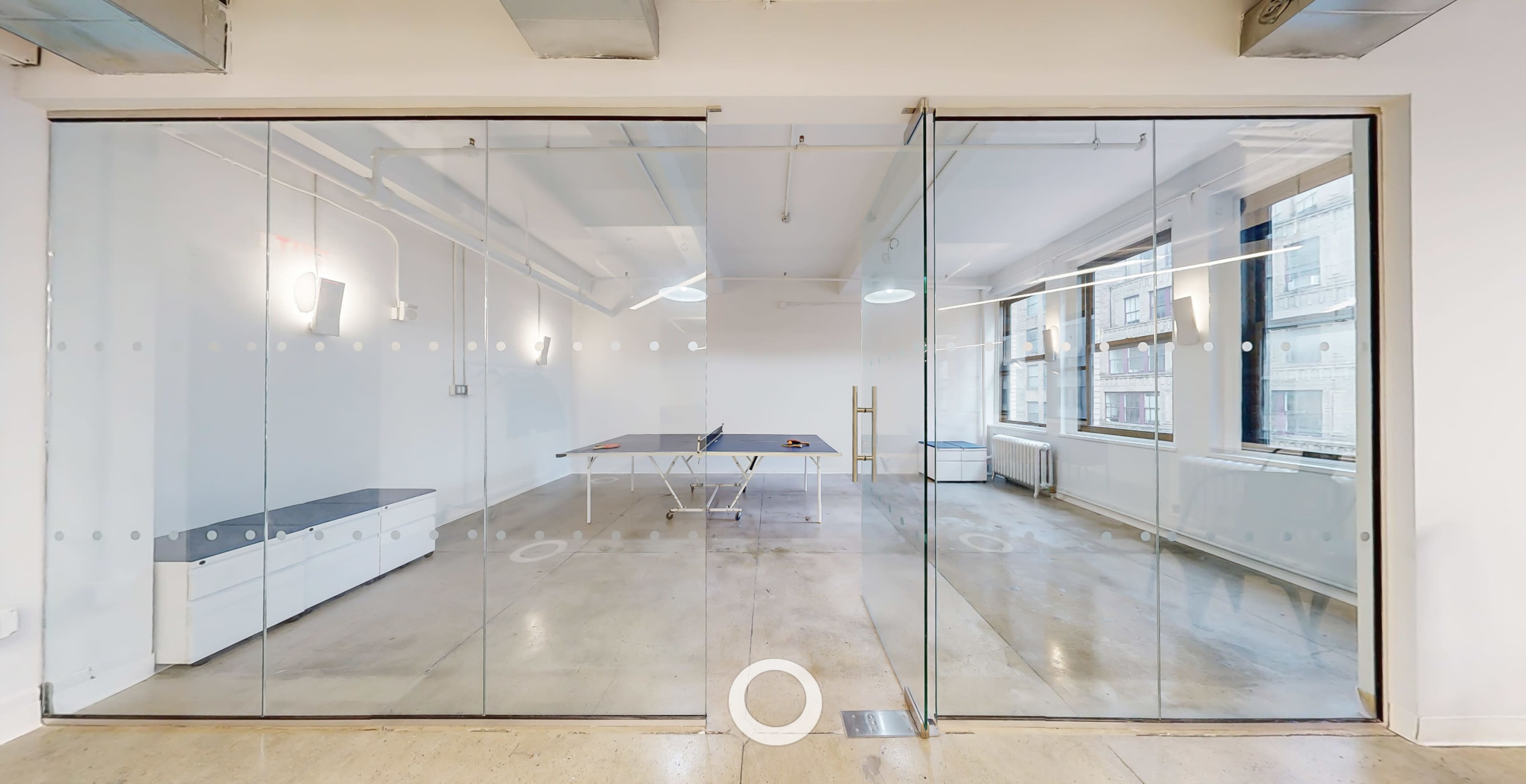 A modern office space with glass walls, featuring a table in the center and large windows allowing natural light to enter.