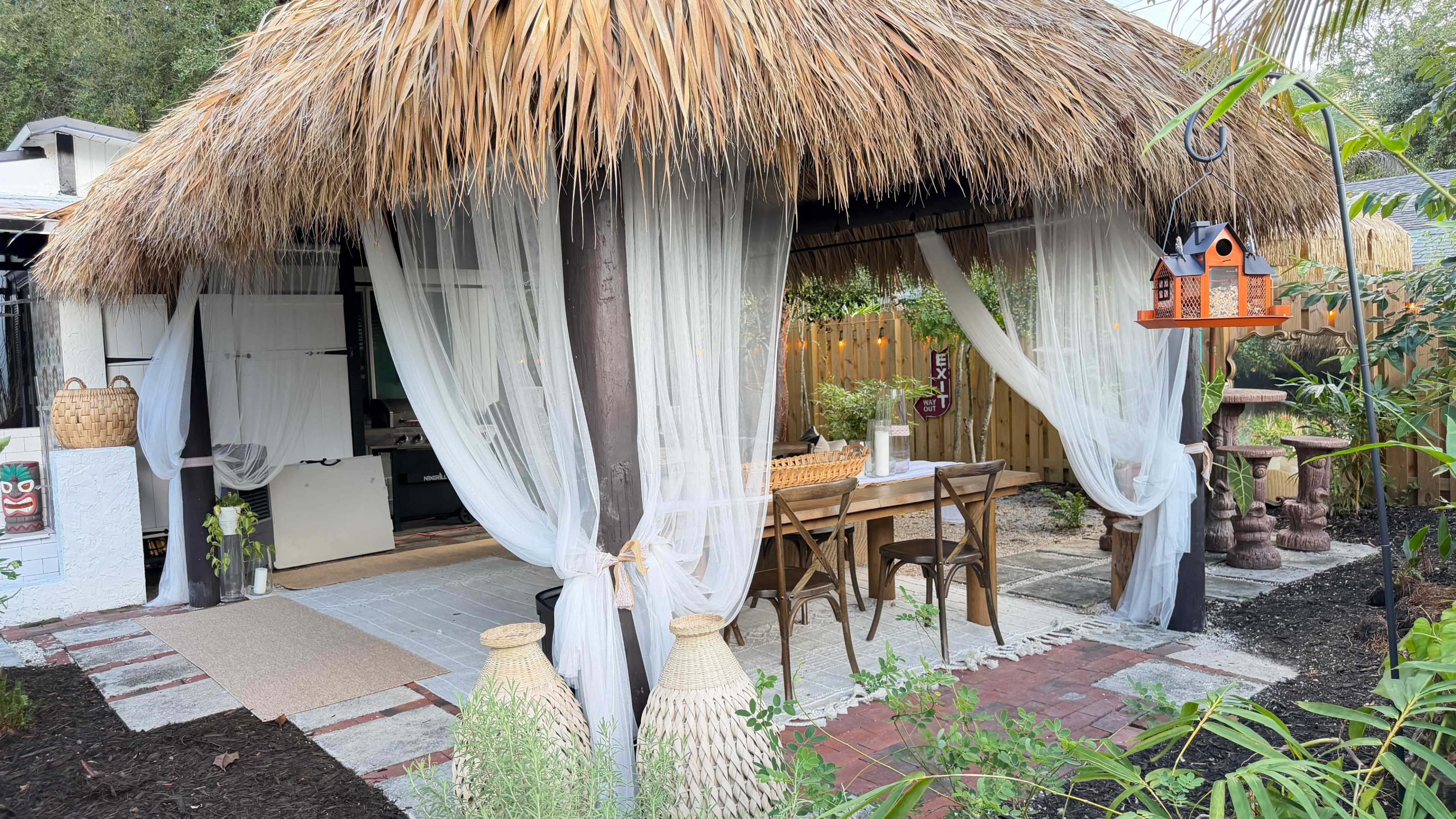 The image shows a thatched-roof outdoor dining area with a table and chairs, surrounded by lush greenery and decorative elements.