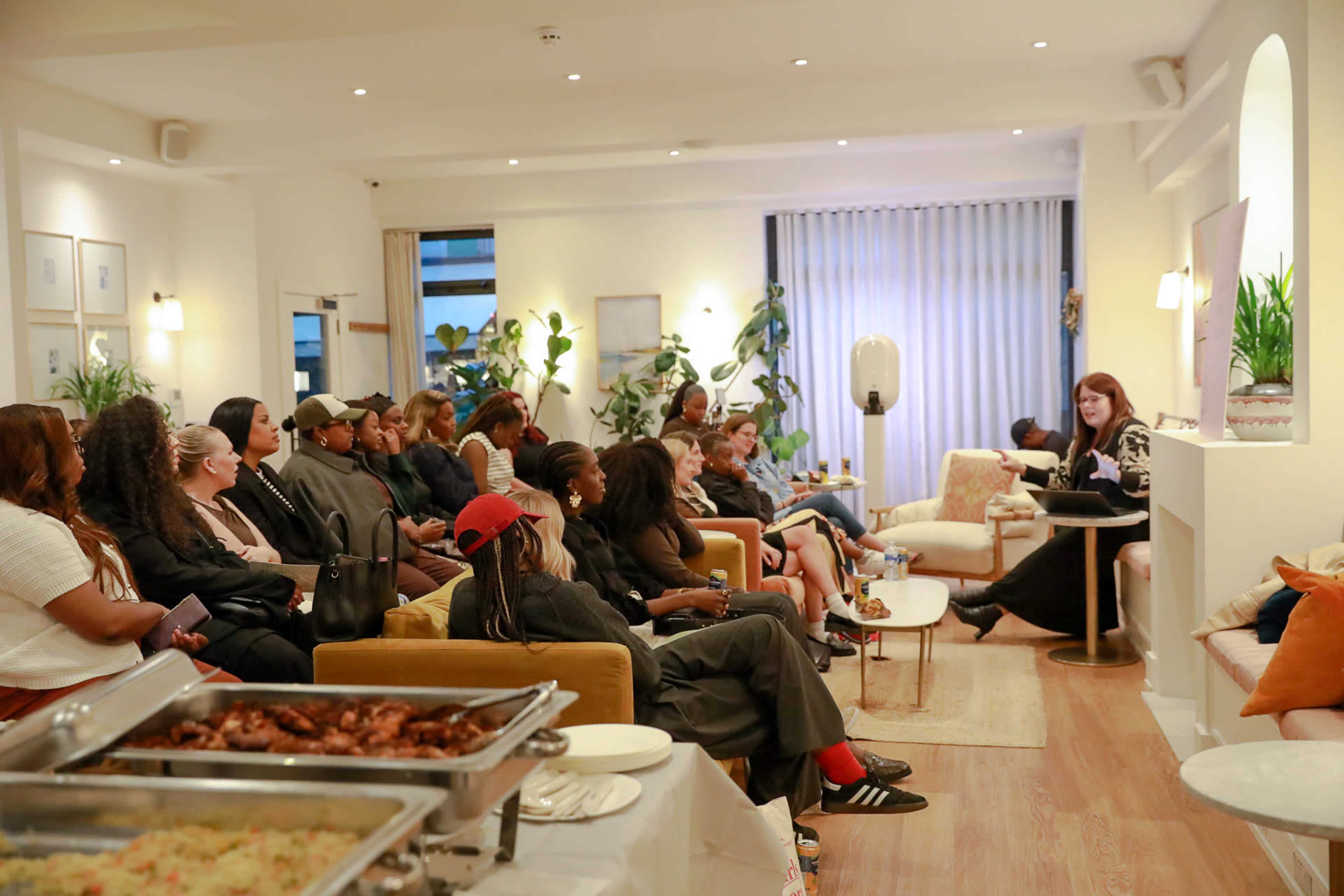 A diverse group of people is seated in a well-lit modern room, listening intently to a woman speaking at the front.