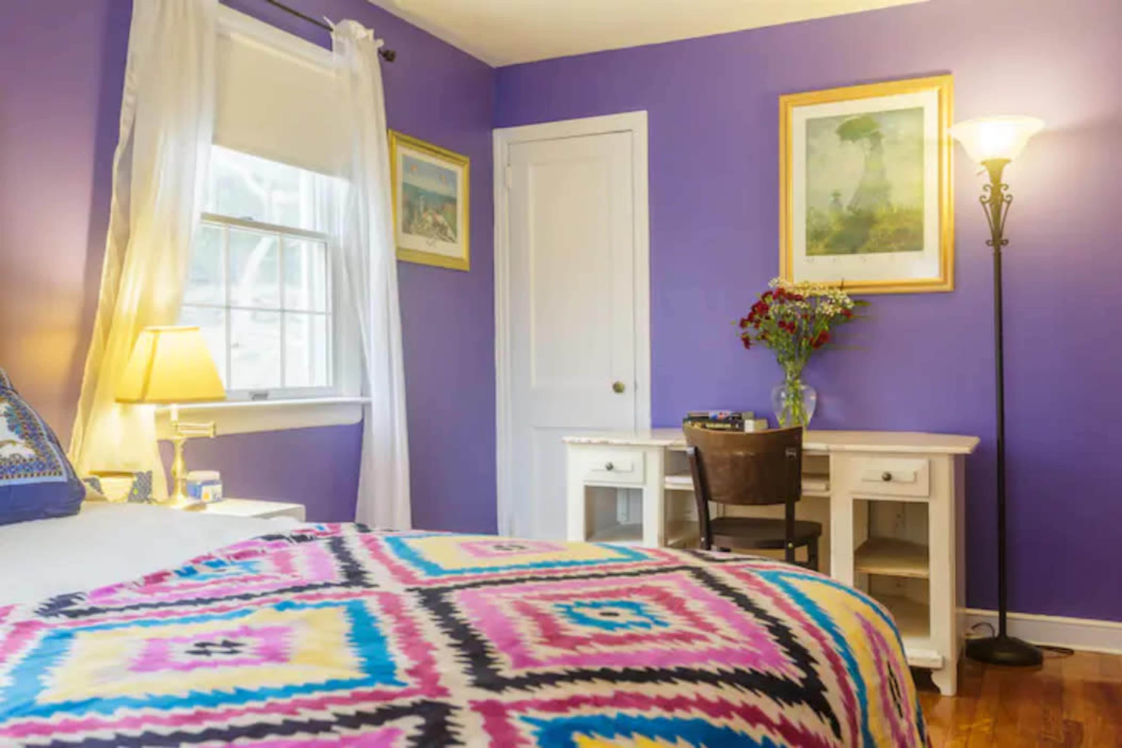 A bedroom with purple walls, a bed with a colorful patterned blanket, a desk with a chair, and a lamp beside the bed.