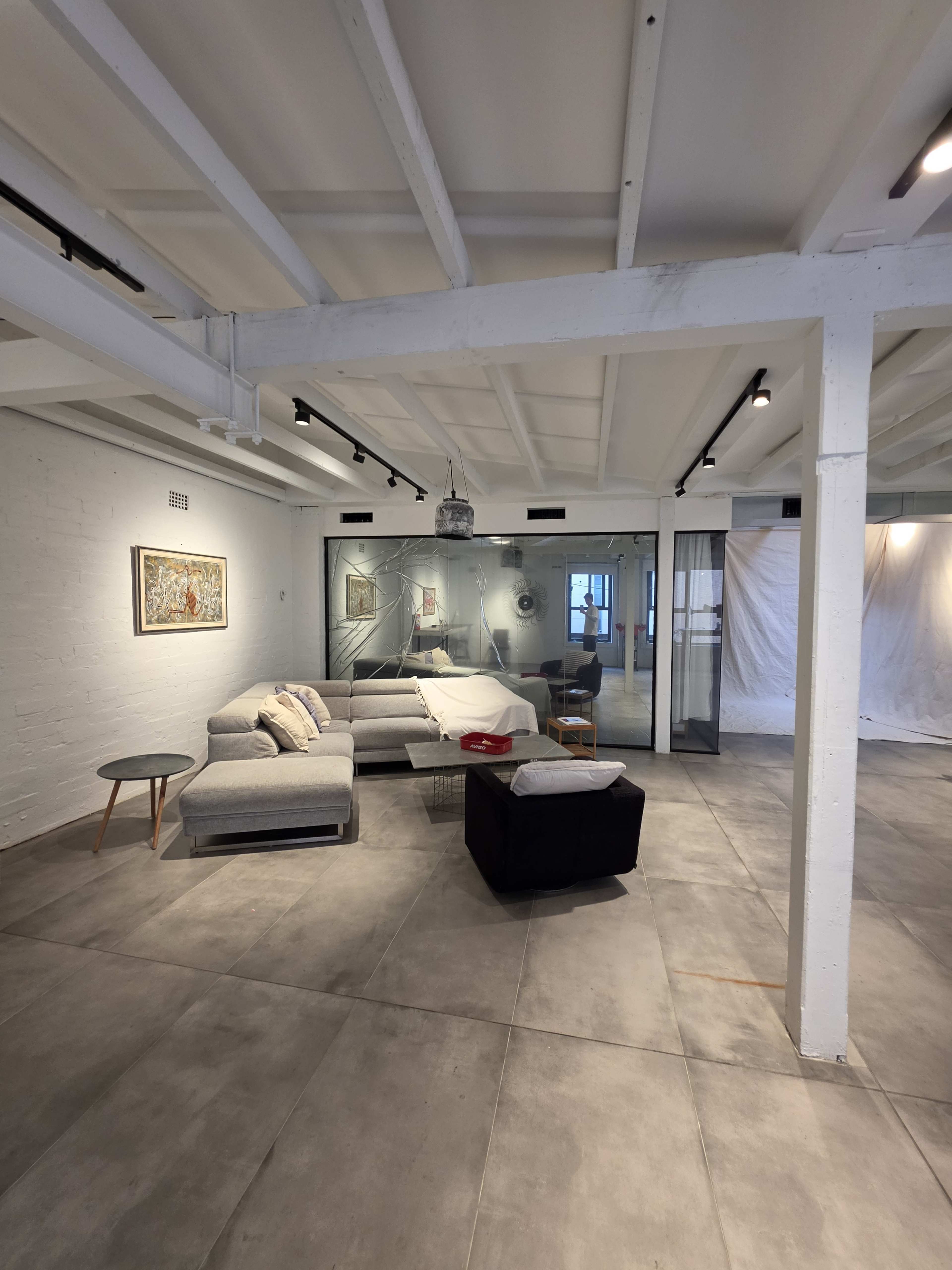 The image shows a spacious, modern living area with a sectional sofa, a coffee table, and a partially glass-enclosed workspace in the background.