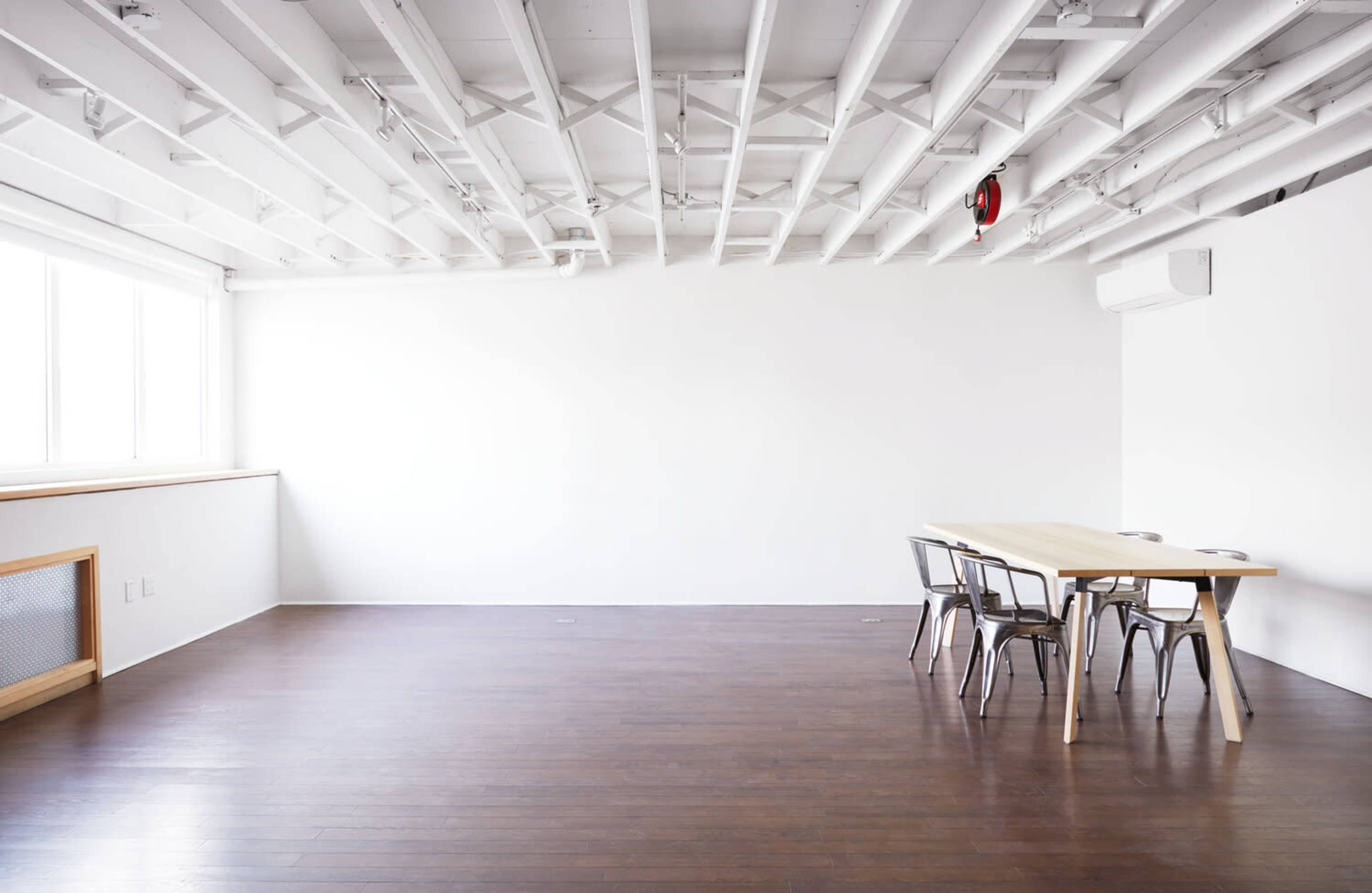 A spacious, empty room features a large wooden table surrounded by metal chairs under a white, exposed beam ceiling.