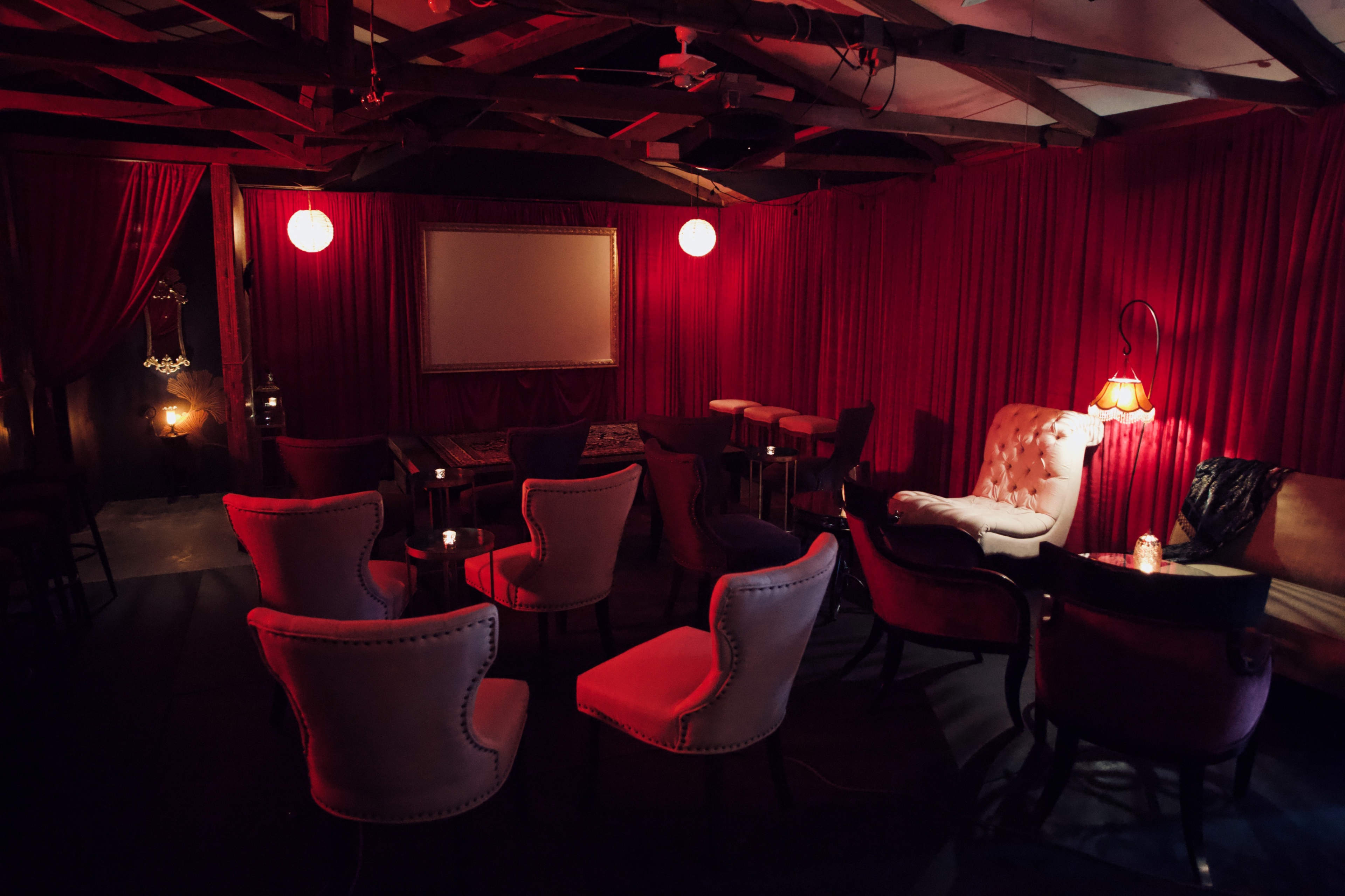 The interior of a dimly lit lounge features plush seating arranged around small tables, with red curtains and soft, ambient lighting.