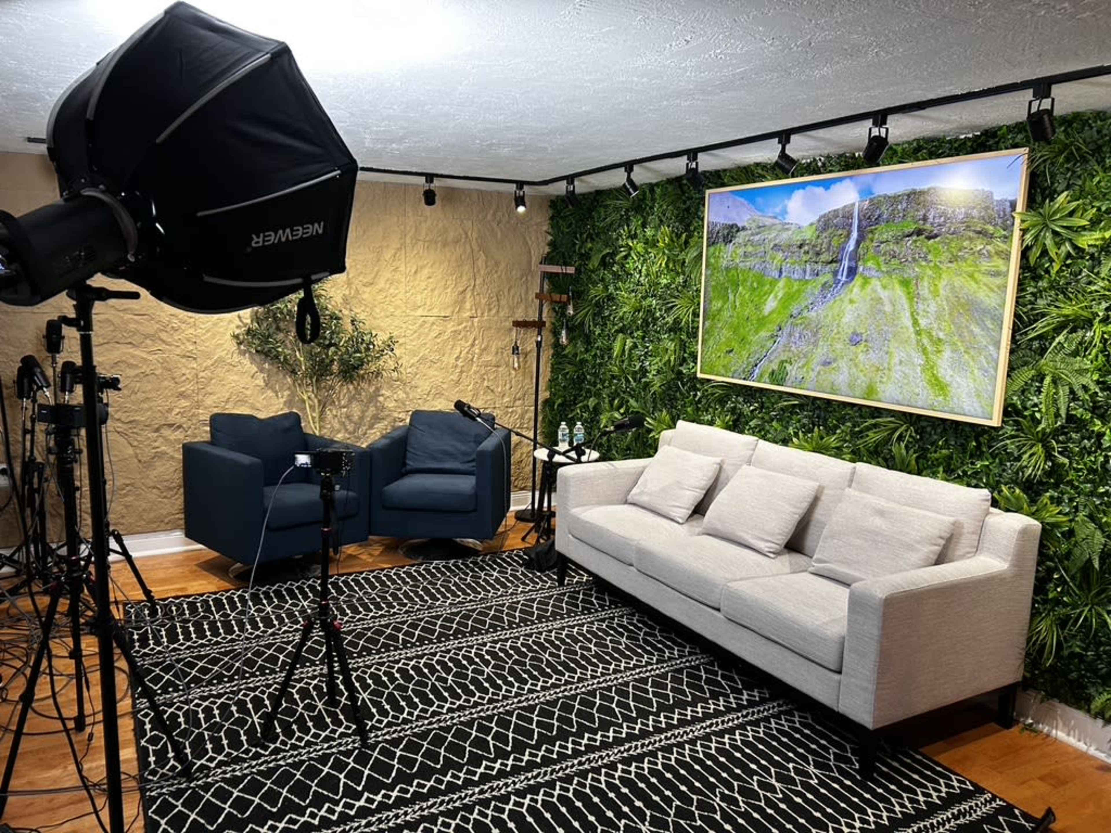 A room set up for filming, featuring a light source, a couch, two blue chairs, and a large landscape photograph on an accent wall covered with greenery.