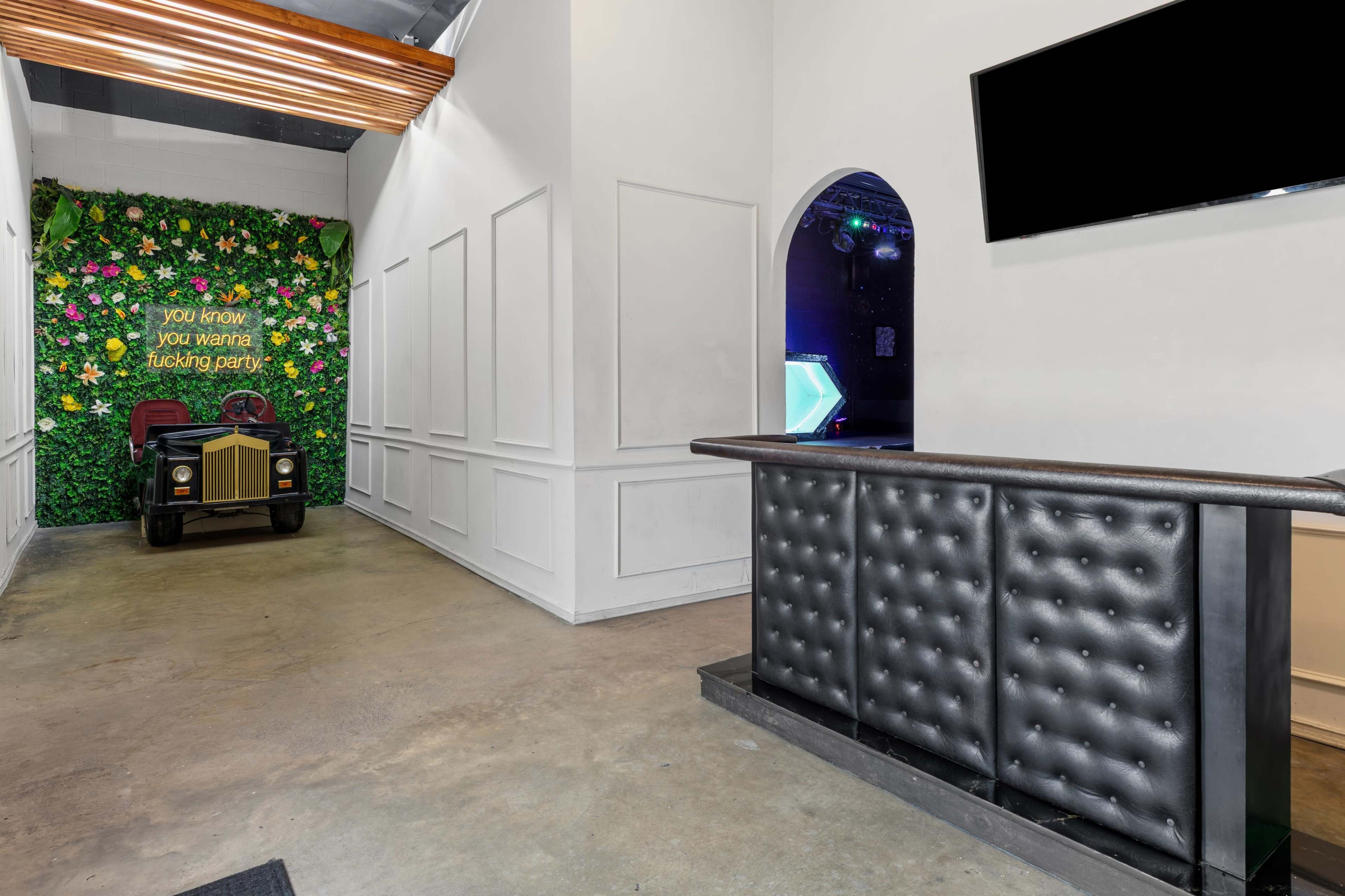 The image shows a hallway with a green floral wall on one side, a reception desk with a tufted black surface, and a vintage-style car positioned nearby.