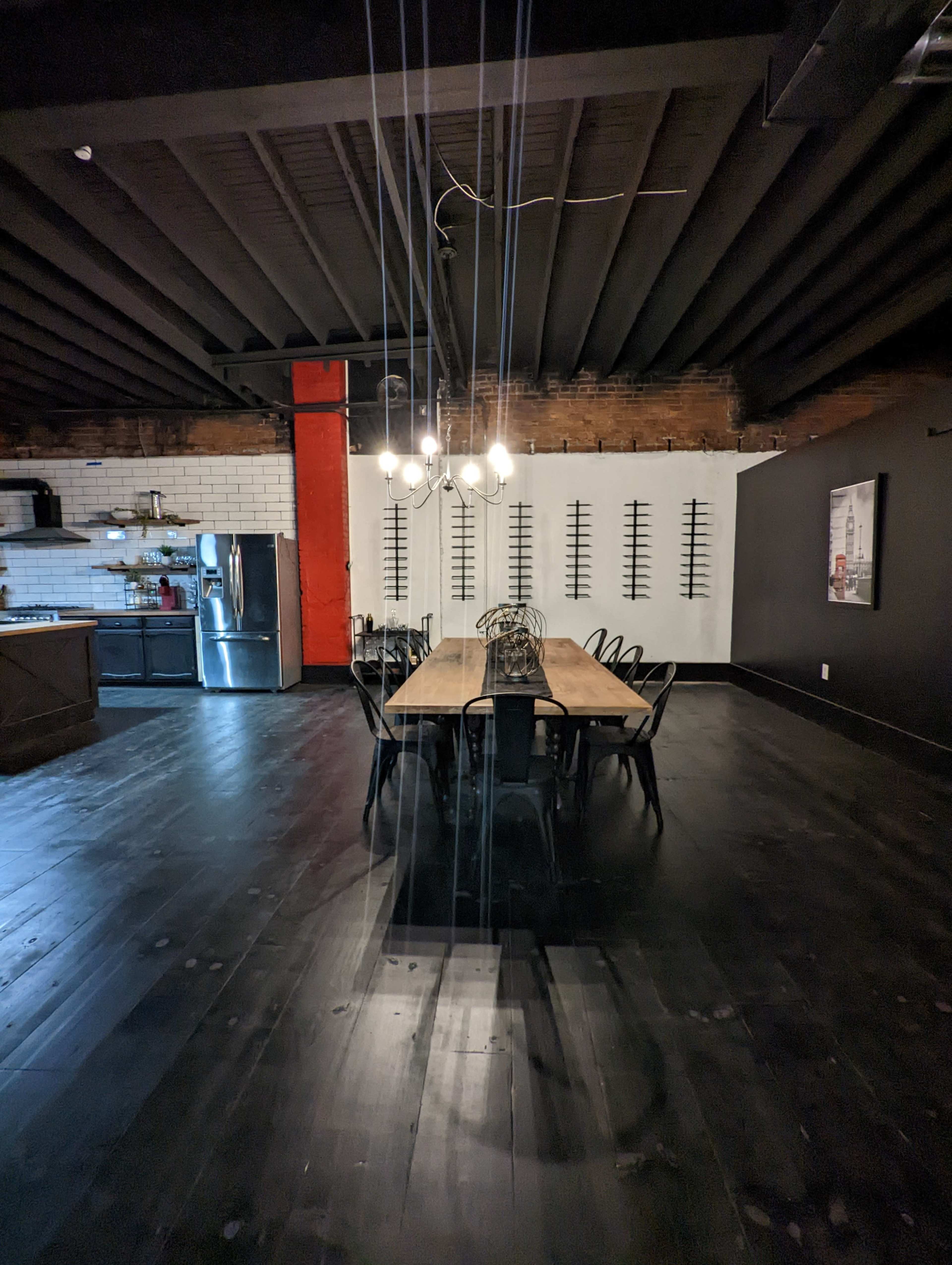 A long wooden dining table with black chairs is set in a spacious room featuring exposed brick walls and a modern kitchen area.