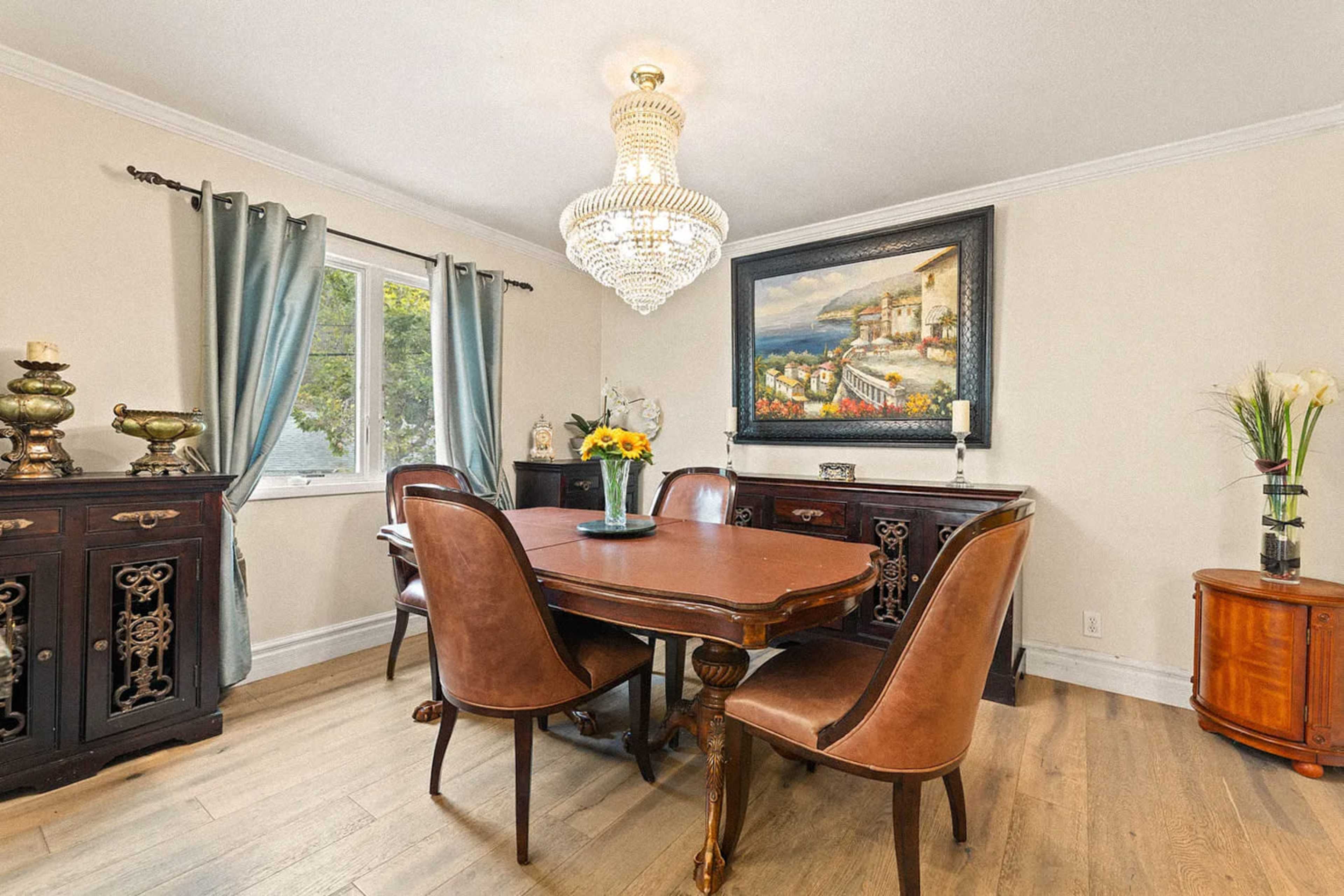 The image shows a dining room with a wooden table surrounded by brown upholstered chairs, a large chandelier overhead, and a decorative painting on the wall.