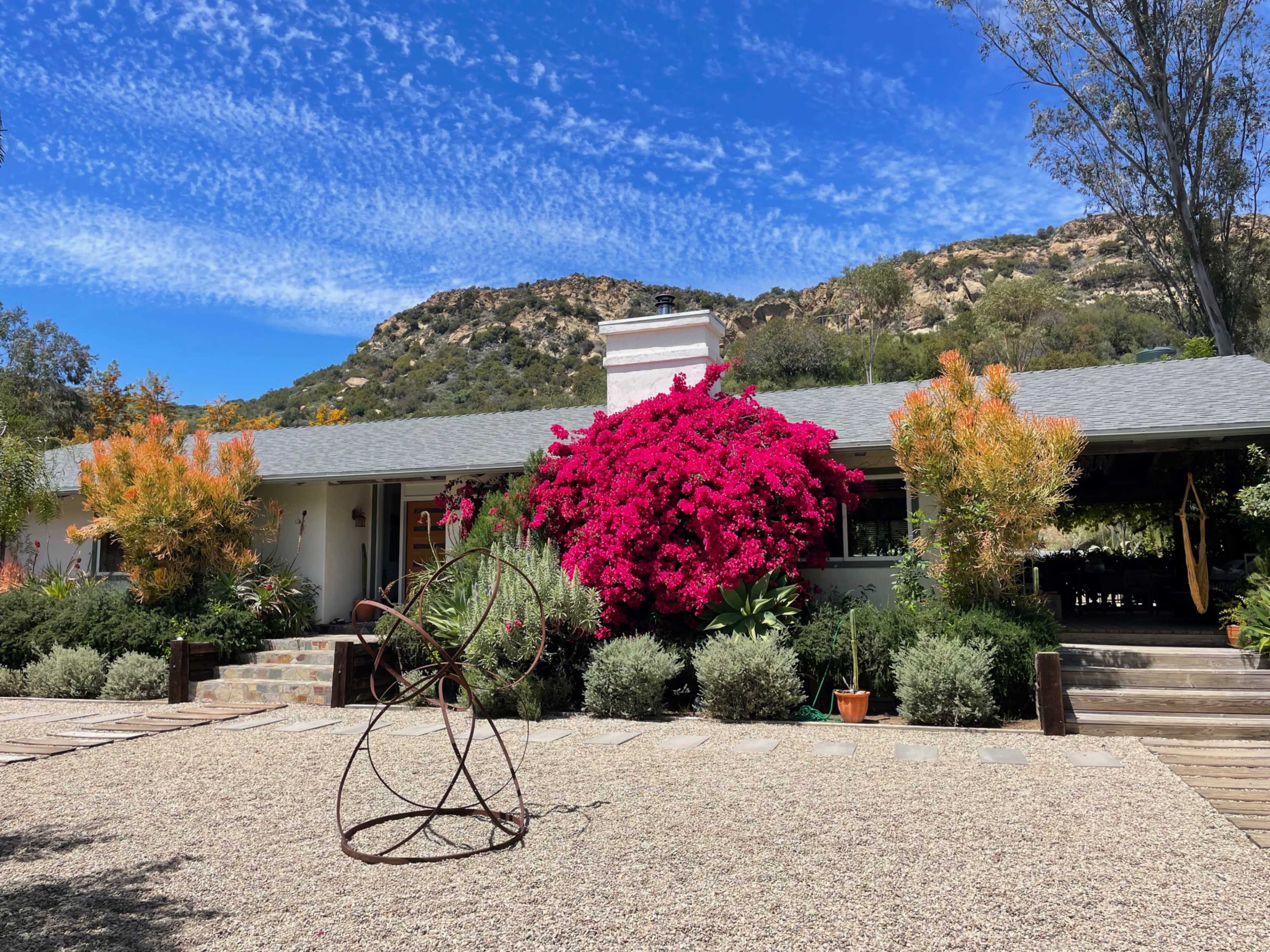A single-story house with a vibrant bougainvillea bush and a gravel driveway is set against a backdrop of hills and a blue sky.