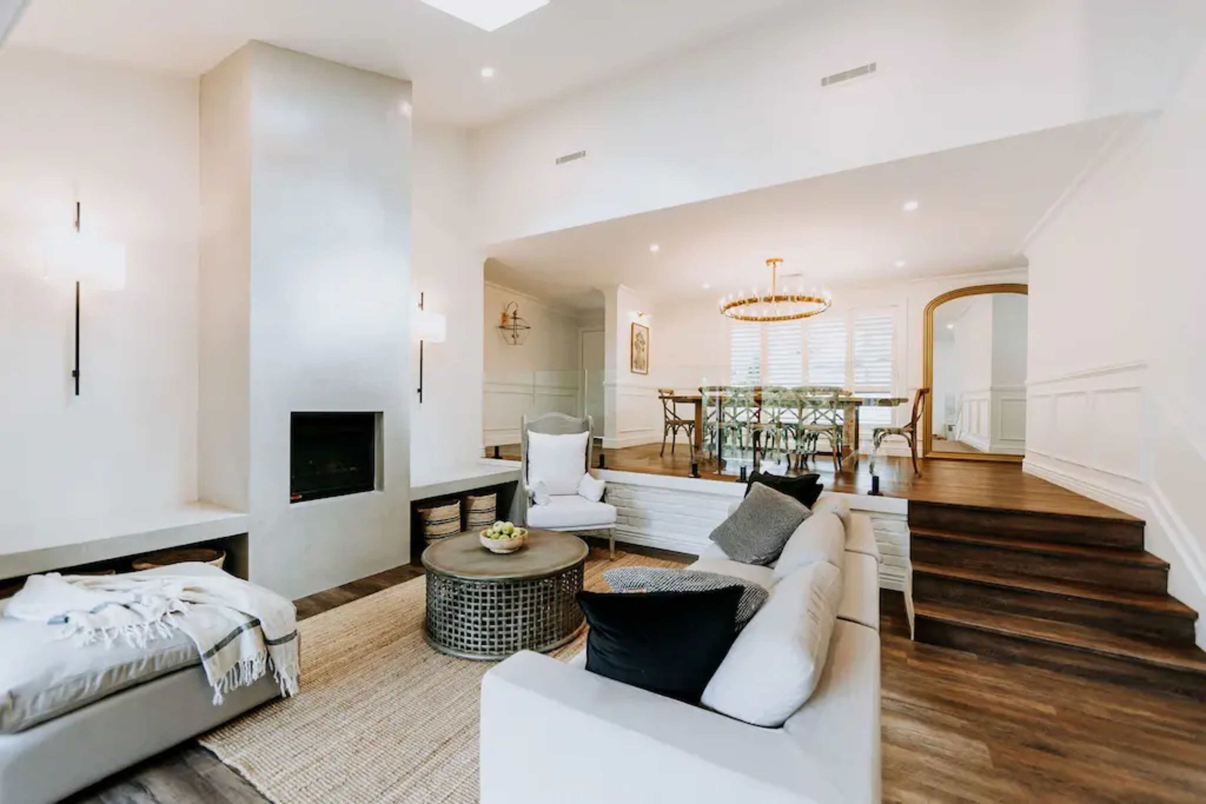 A modern living room with a light-colored sofa, a circular coffee table, a fireplace, and dining area visible in the background with a large chandelier.