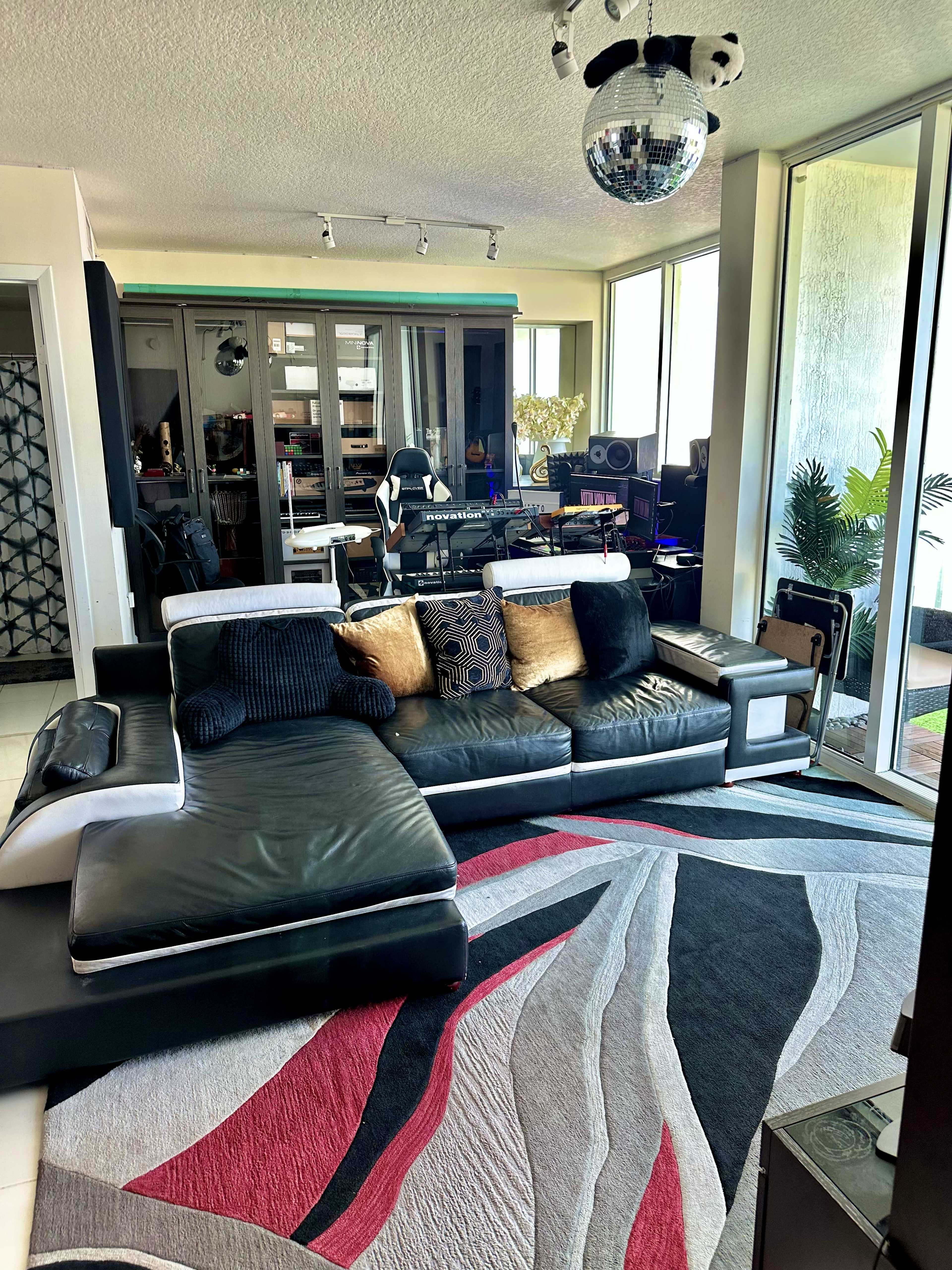 A modern living room with a large black sectional sofa, a patterned area rug, and a workspace featuring music equipment in the background.