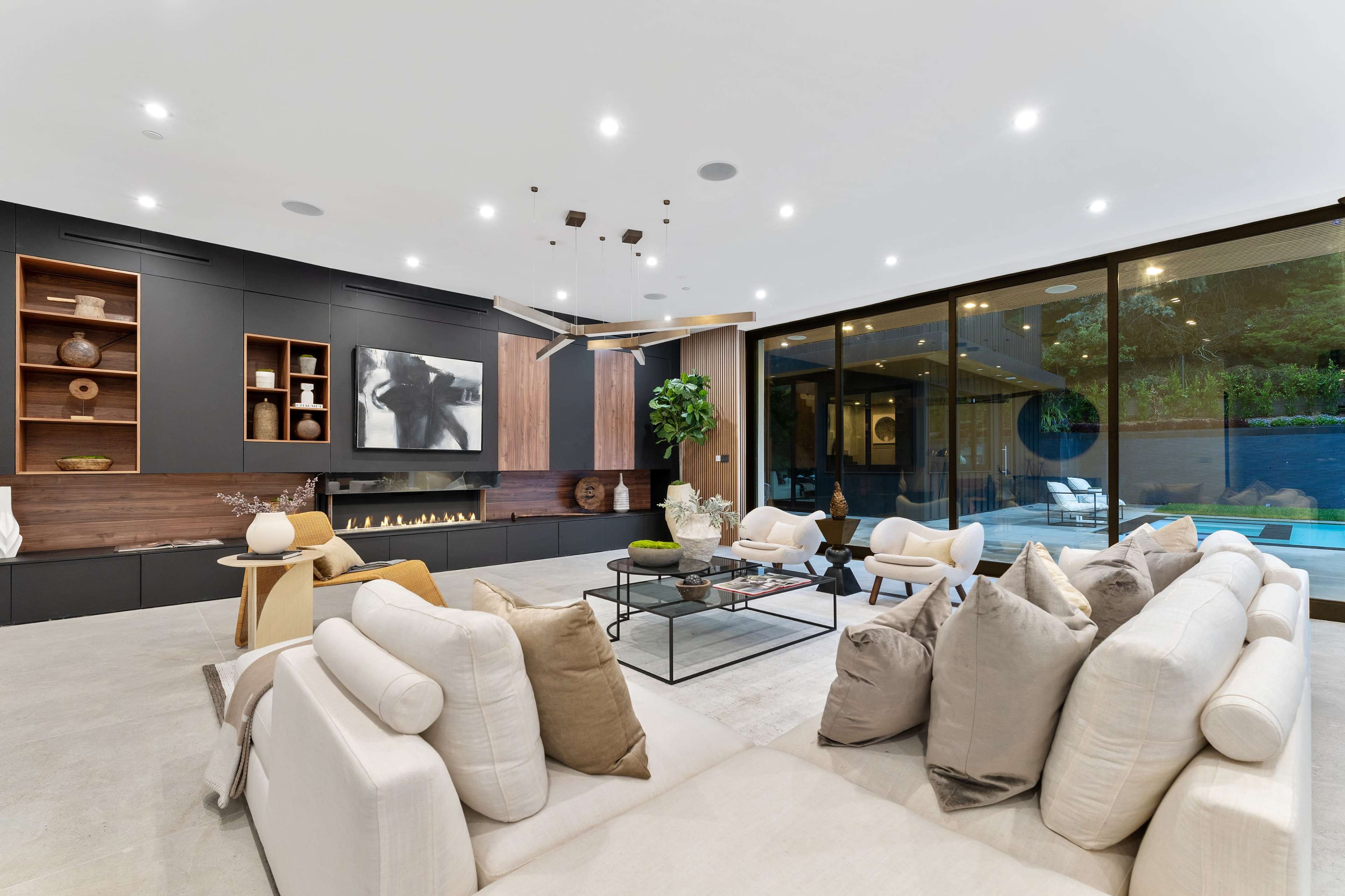The image depicts a modern living room with a large glass wall overlooking an outdoor space, featuring a minimalist design, a sectional sofa, and a decorative coffee table.