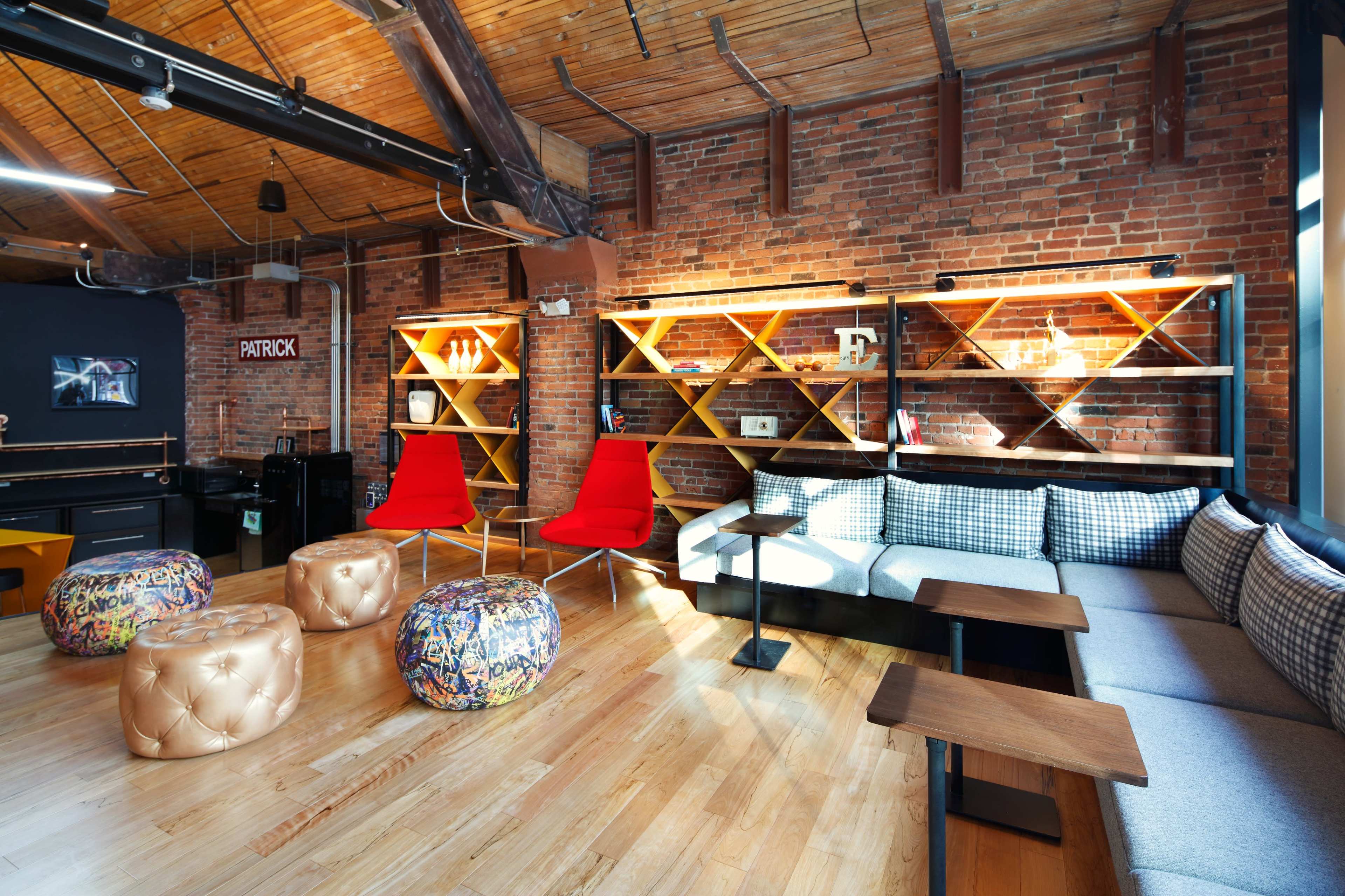 A modern workspace features wooden floors, exposed brick walls, and various seating options, including red chairs and colorful stools, along with shelving displaying decorative items.