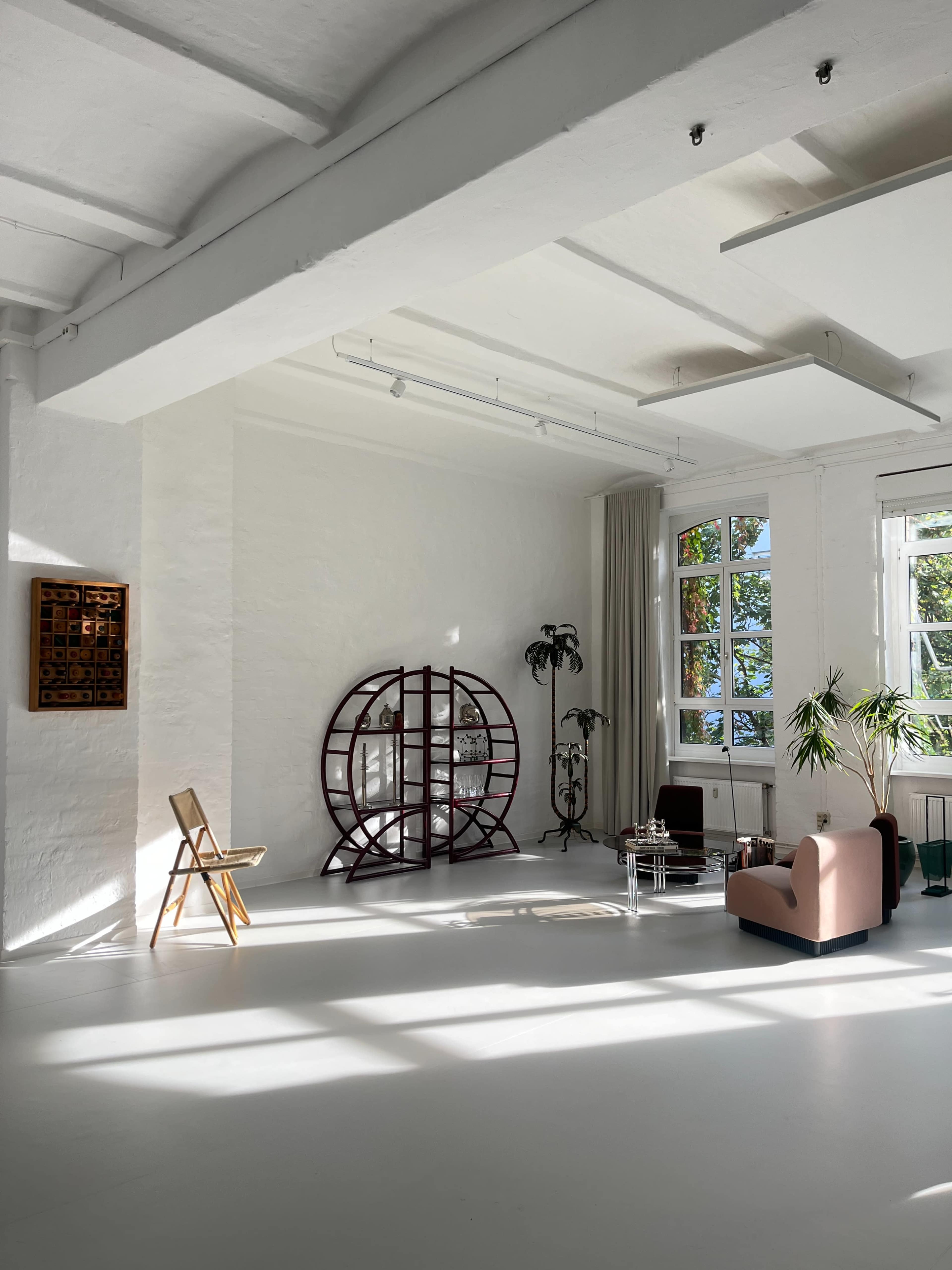 The image depicts a bright, airy living space with large windows, minimalist furniture, and a decorative screen against a white wall.