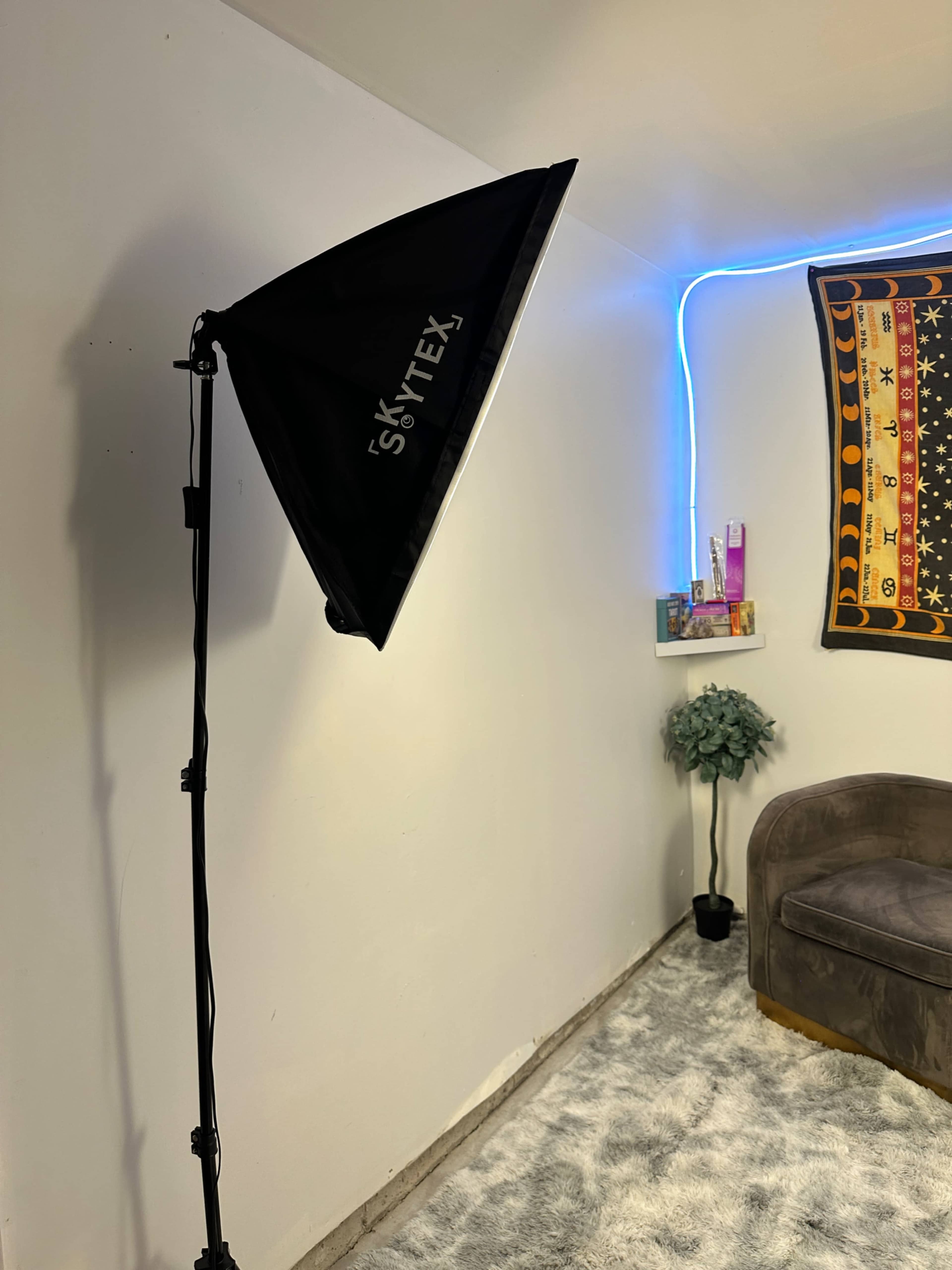 A softbox light stands on a tripod beside a gray couch in a room with a colorful wall tapestry and decorative shelf.