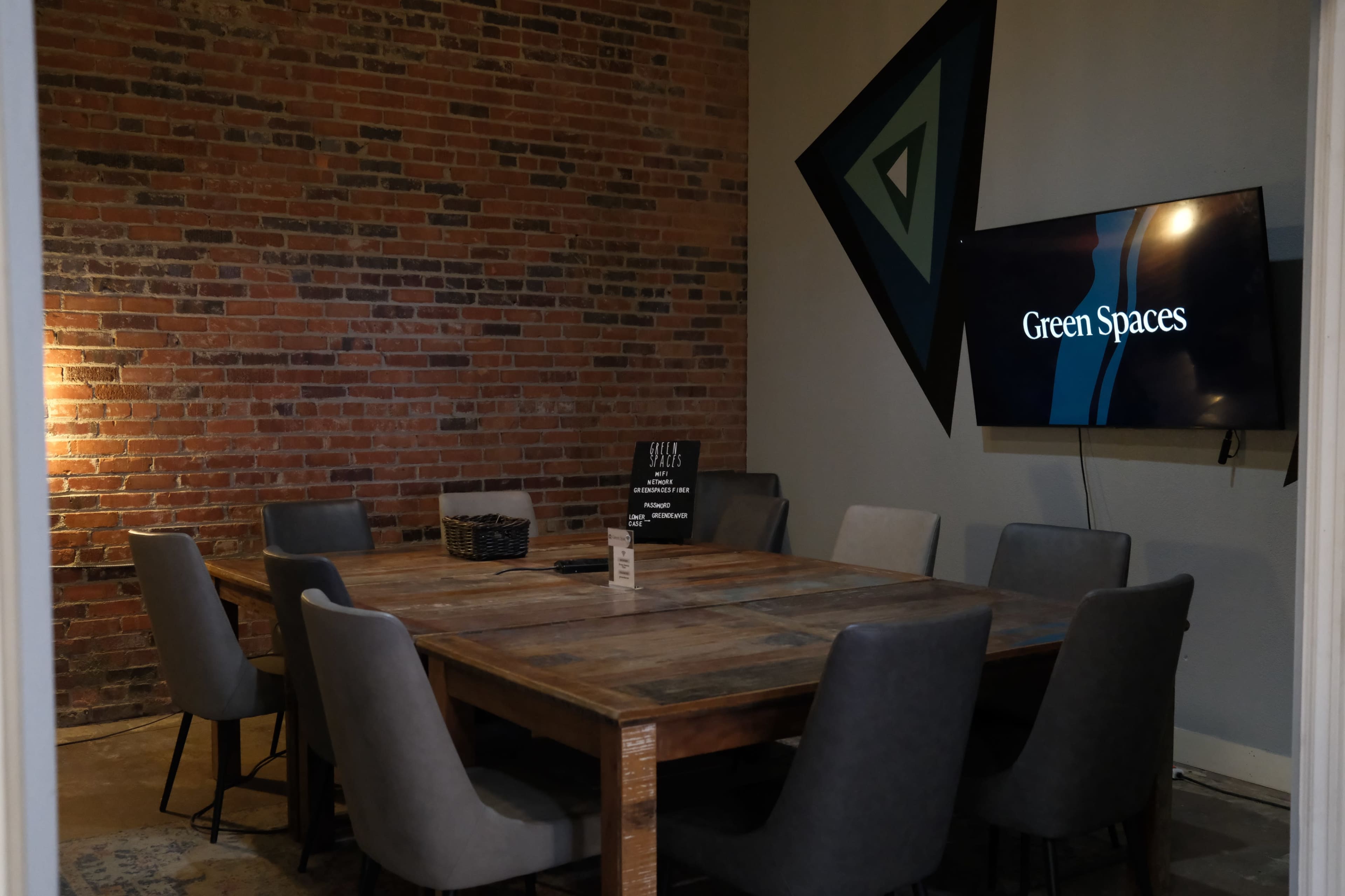 The image shows a conference room with a large wooden table surrounded by gray chairs, a brick wall, and a television displaying the words "Green Spaces."