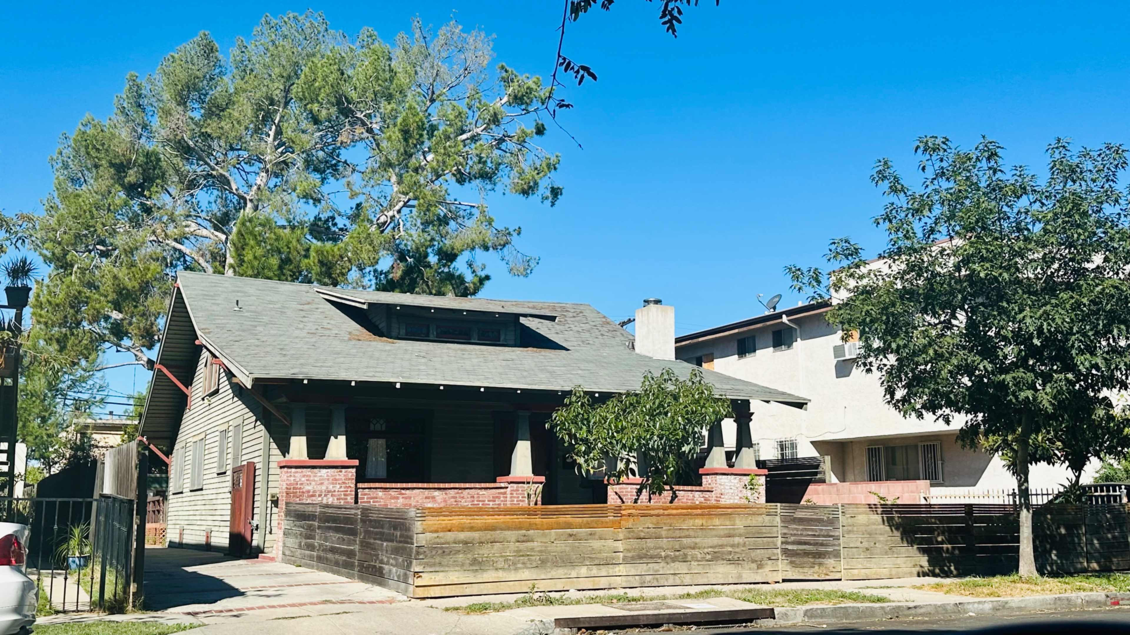 A single-story house with a sloped roof and a front porch is situated next to a larger, multi-story building under a clear blue sky.