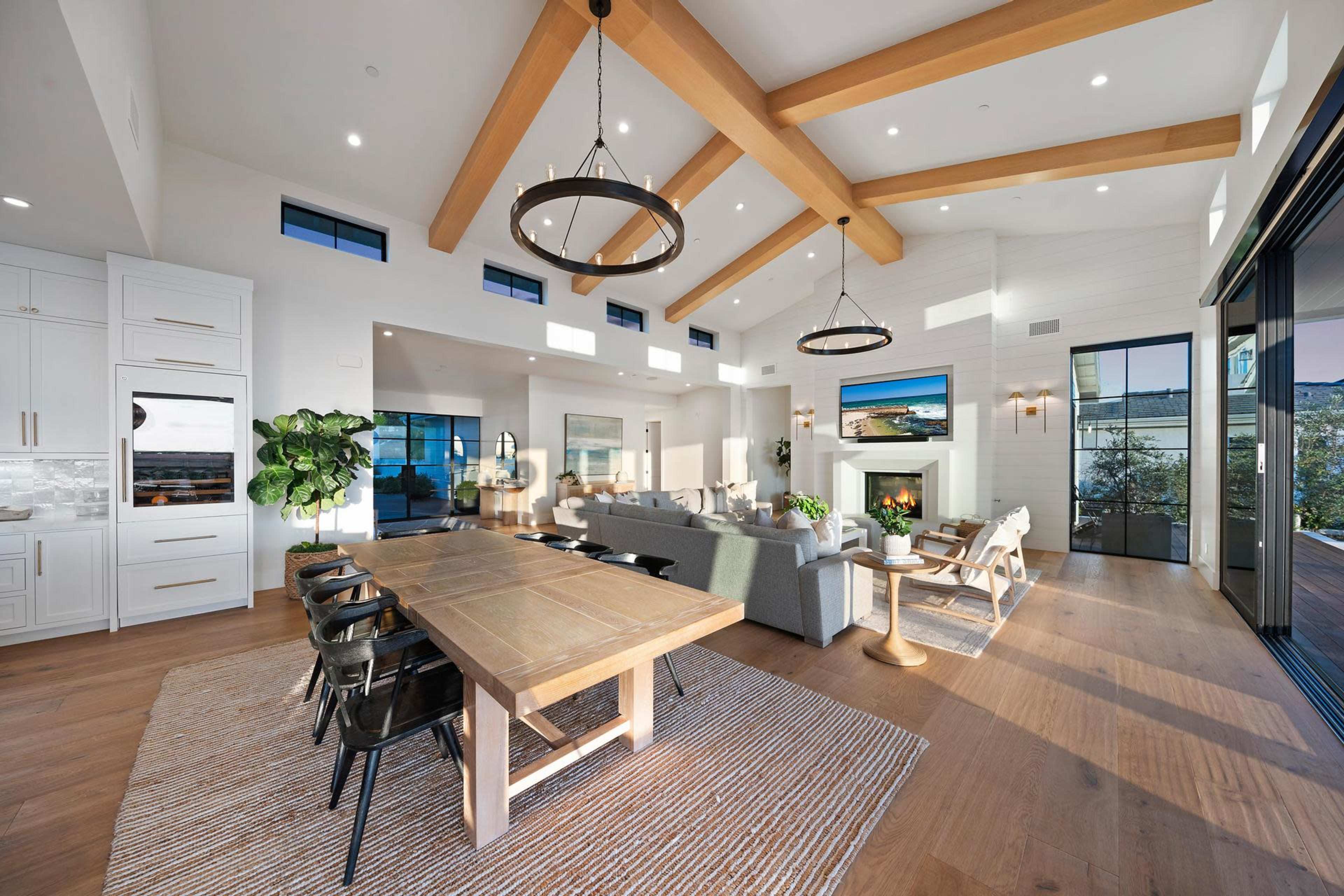 The image shows a bright and modern living space with a high ceiling featuring wooden beams, a large dining table, and a comfortable seating area around a fireplace.