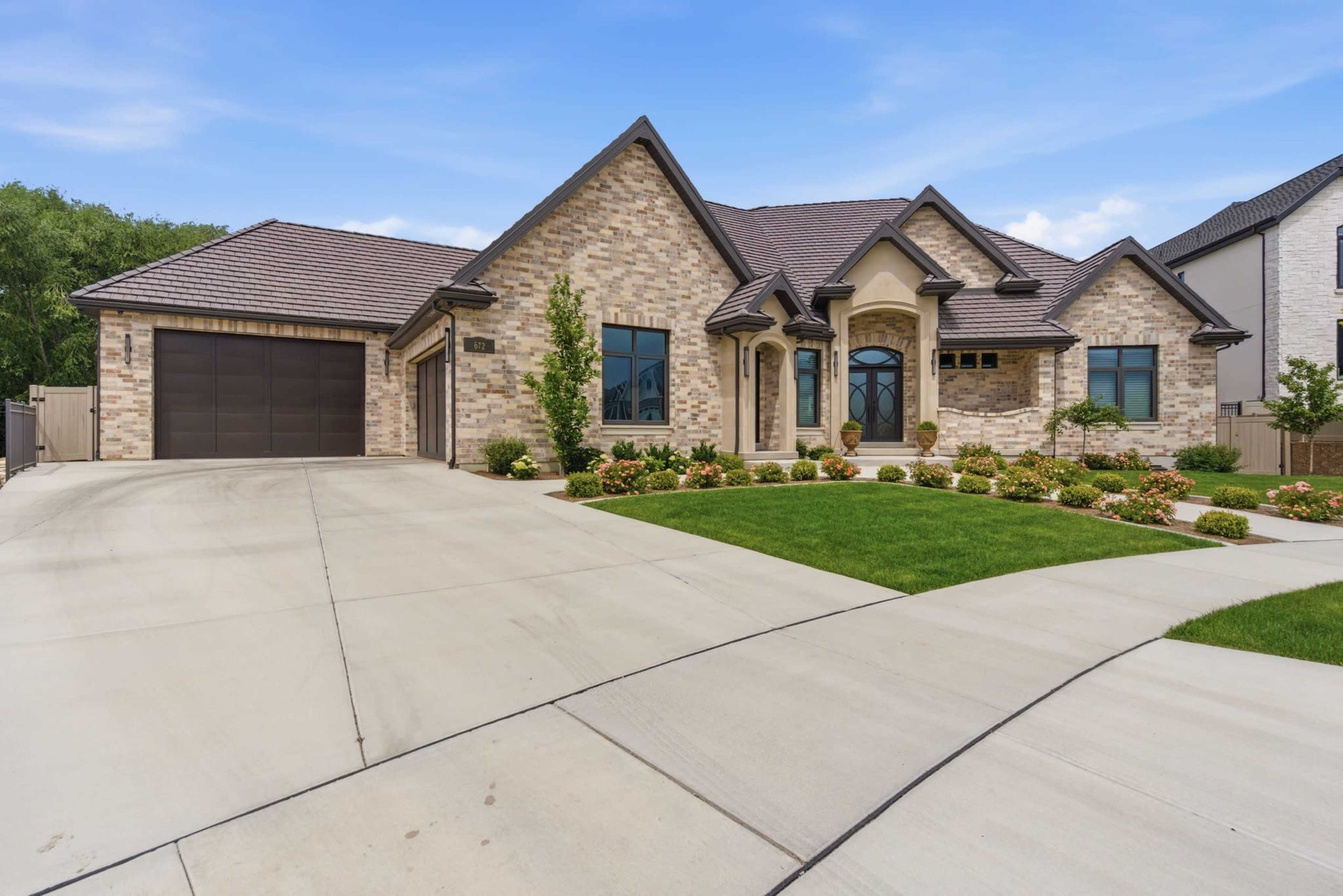 The image shows a large, brick single-story house with a dark roof, a well-manicured front yard, and a concrete driveway leading to a two-car garage.