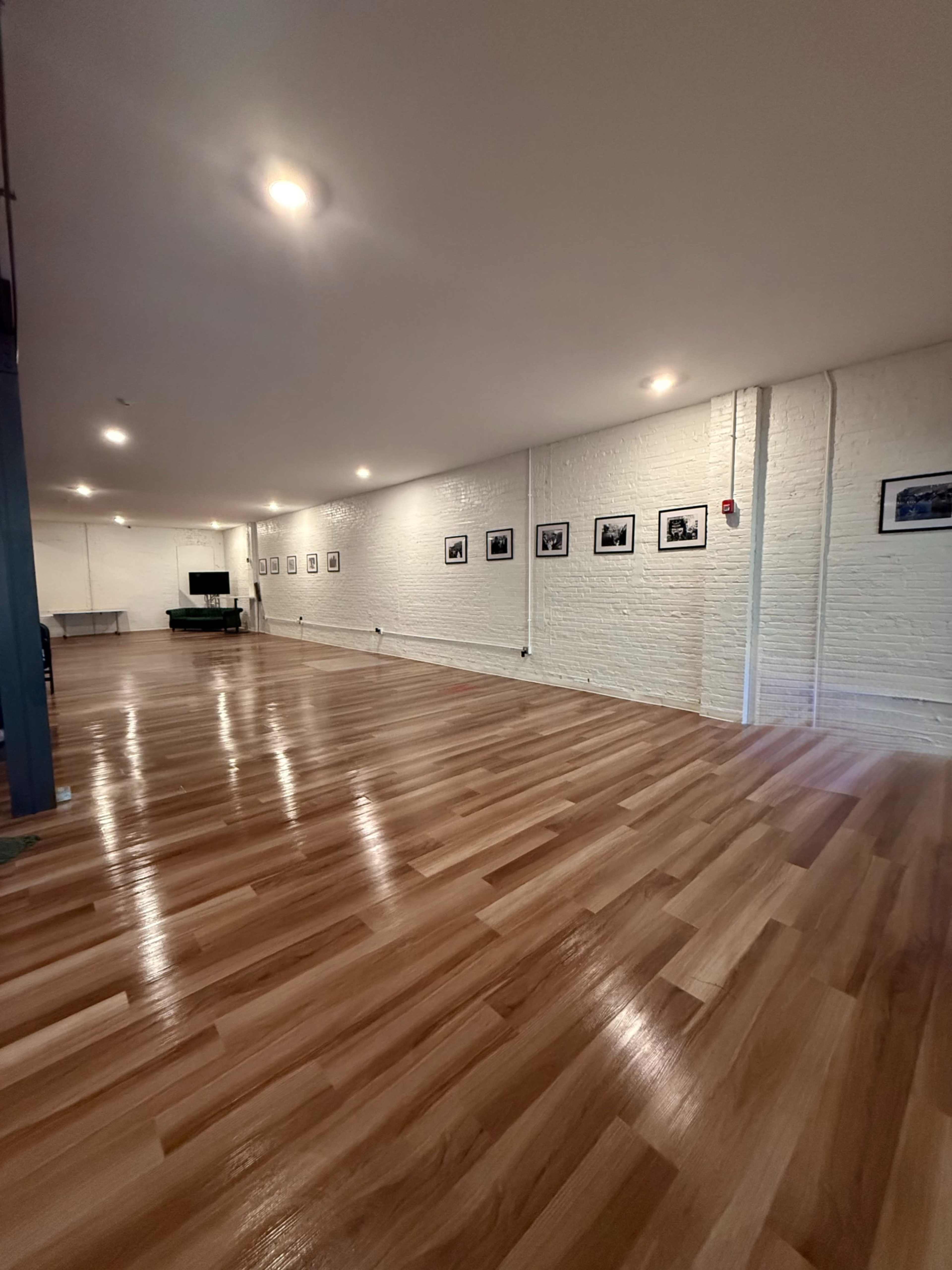 The image shows a spacious interior with polished wooden floors, white brick walls, and several black-and-white photographs hung along one side.