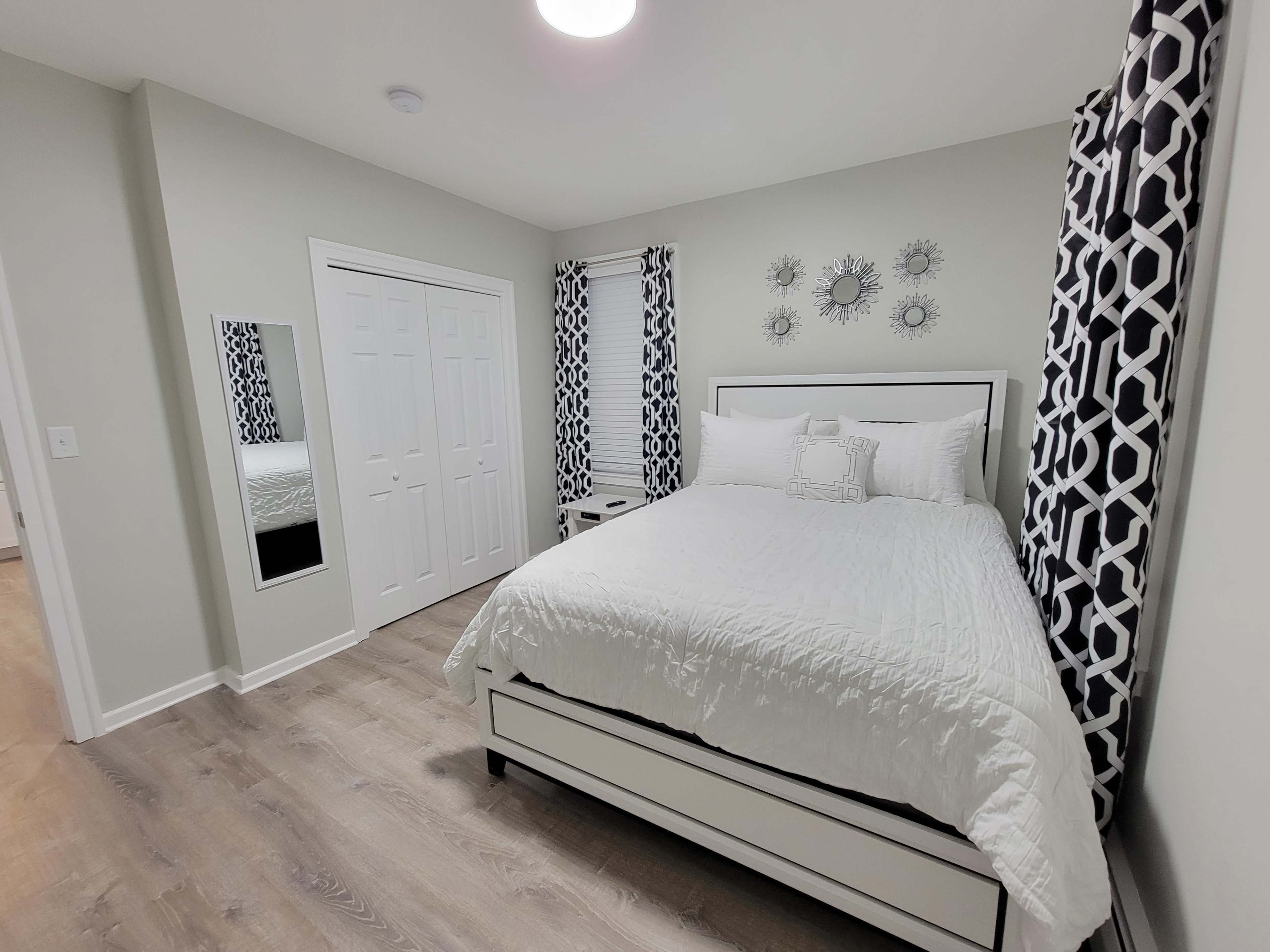 A modern bedroom with a white bed, patterned black and white curtains, and a mirror on the wall.
